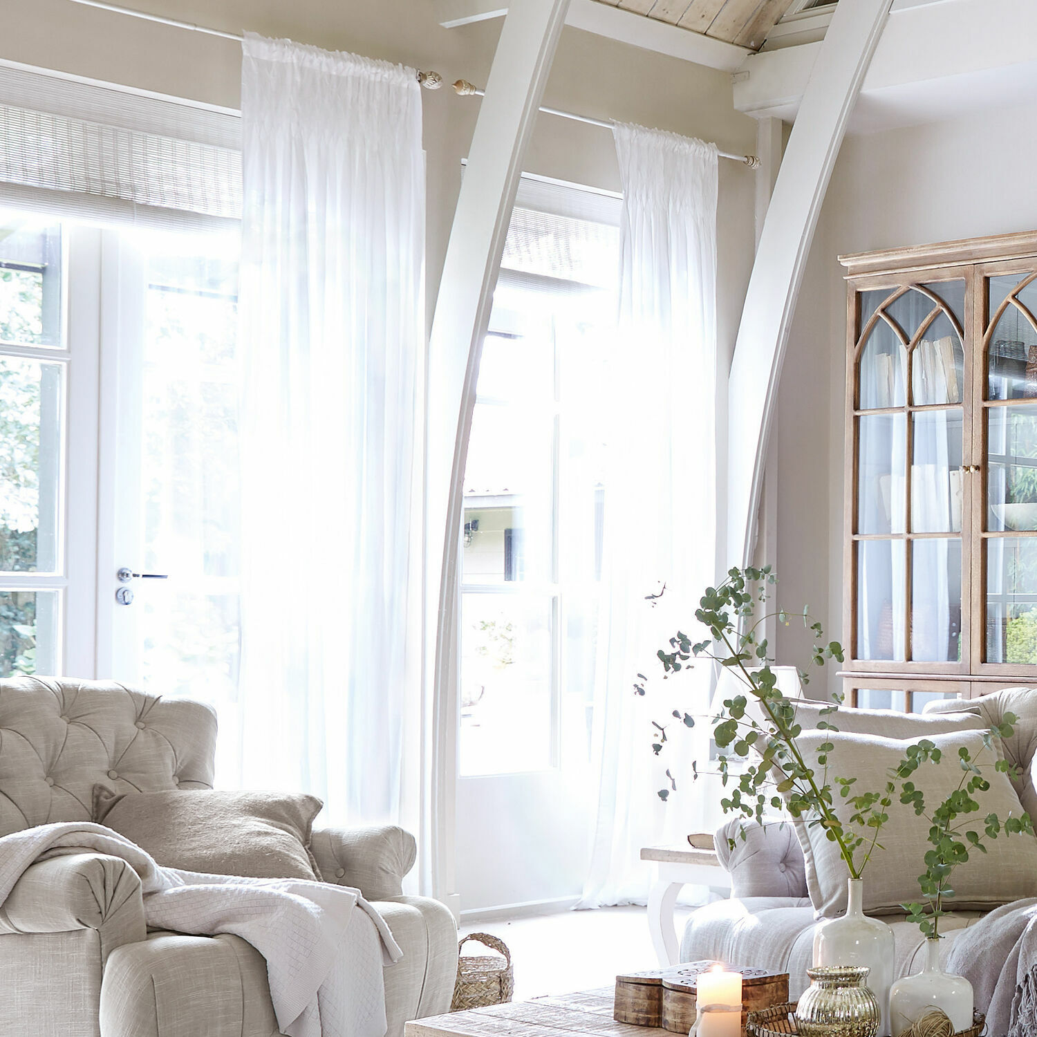 Gardine Otilie Helles Wohnzimmer mit beigem Polstersessel, weissen Vorhängen und Vintage-Holzvitrine