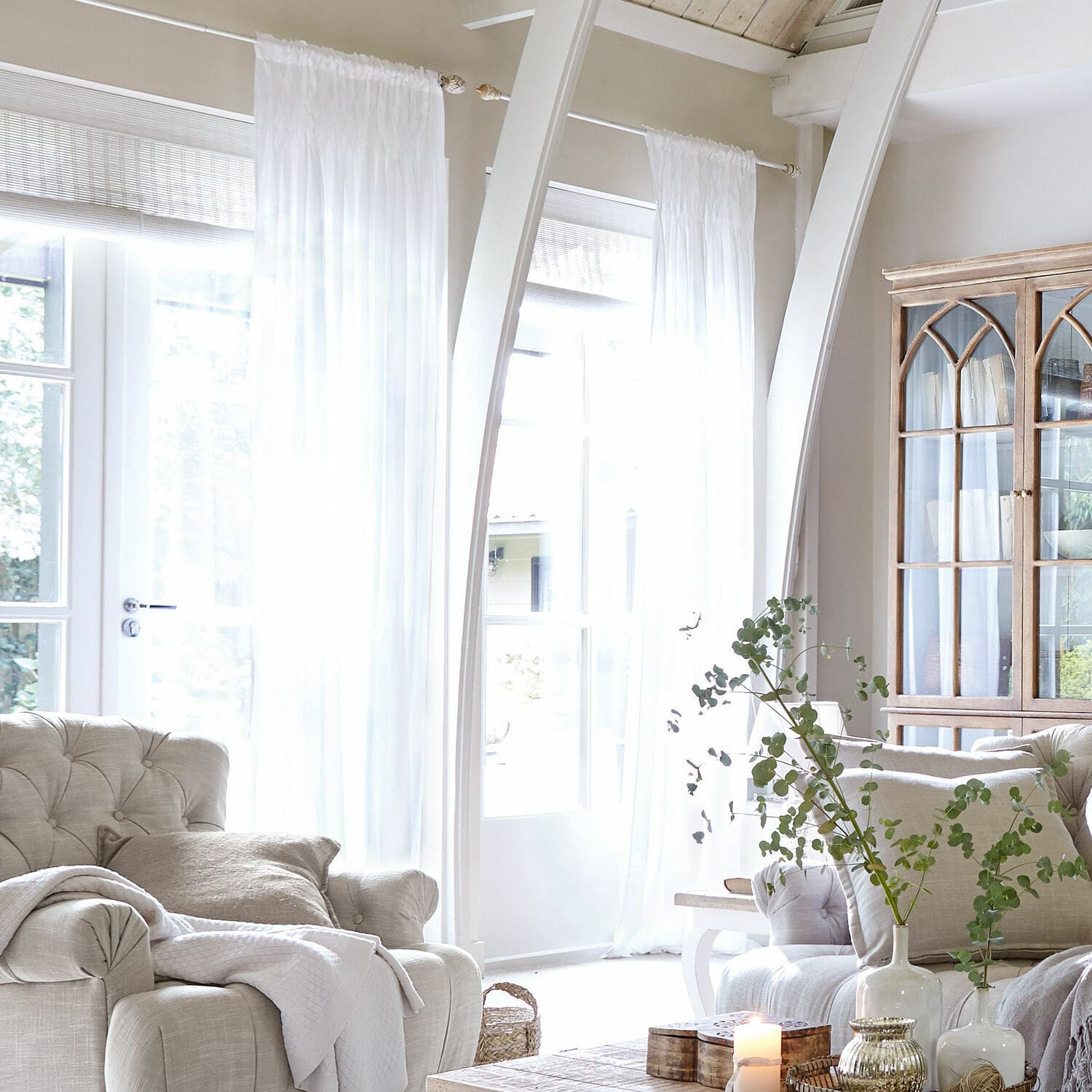 Gardine Otilie Helles Wohnzimmer mit beigem Polstersessel, weissen Vorhängen und Vintage-Holzvitrine