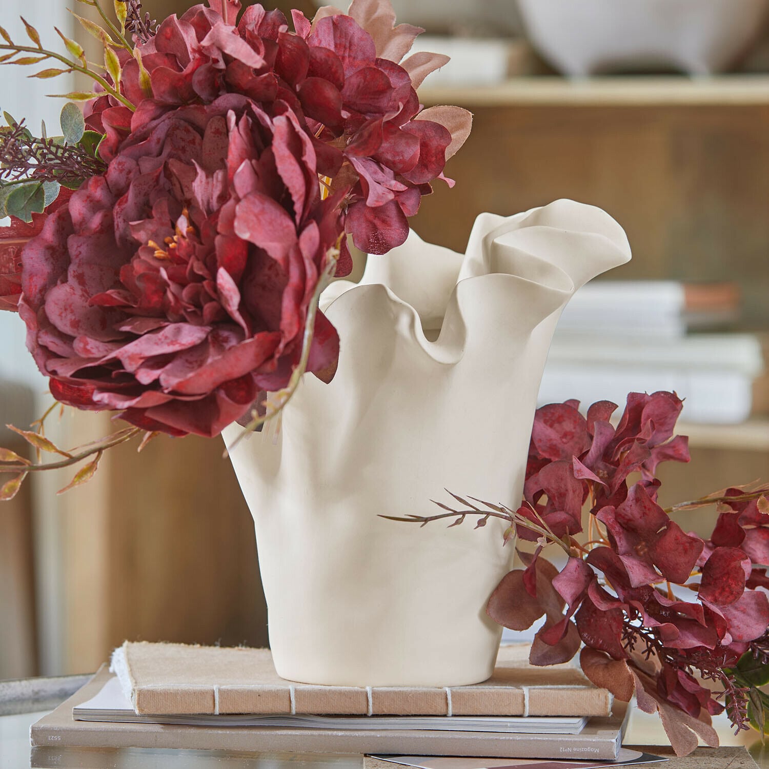 Vase Elvessa Weisse Keramikvase mit wellenförmigem Rand und roten künstlichen Blumen auf Tisch mit Büchern