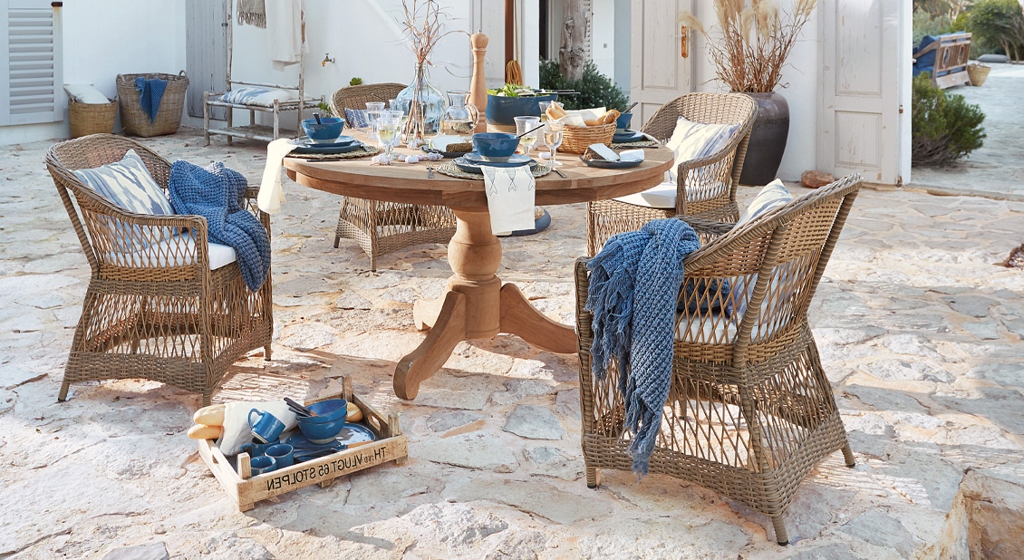 Runder Holztisch mit Rattanstühlen und blauen Accessoires auf steinernem Patio