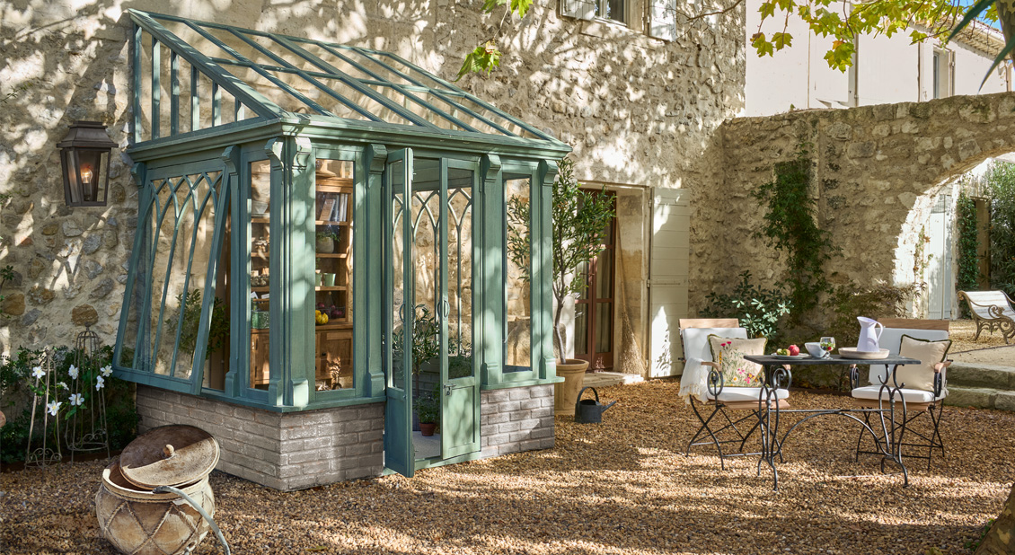 Grünes Metall-Gewächshaus auf Steinsockel in sonnigem Innenhof mit Gartenmöbeln aus Schmiedeeisen