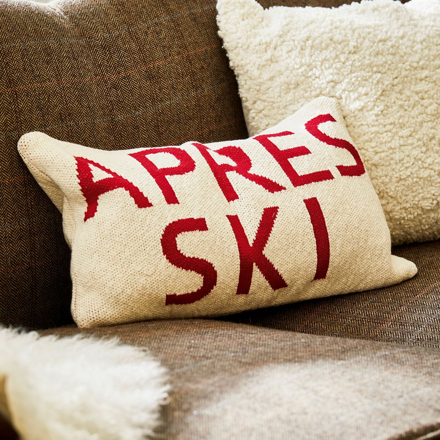 Kissenhülle Apres Ski Beiges Kissen mit rotem Schriftzug Apres Ski auf braunem Sofa mit weichem Fellplaid.