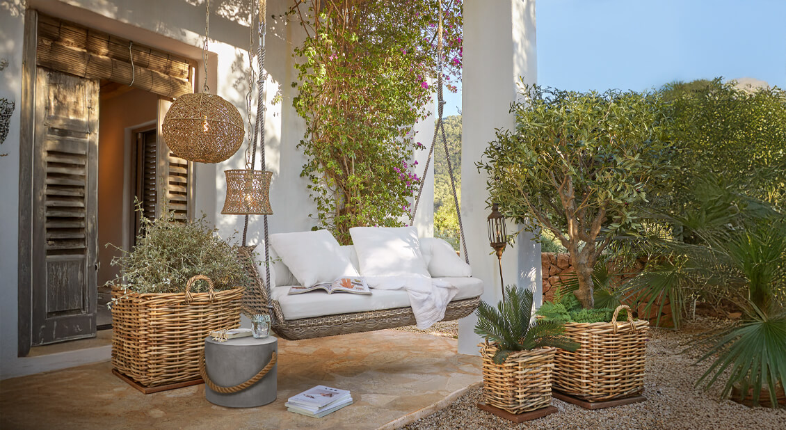 Wiklingschaukel mit weissen Kissen auf Terrasse mit Rattanlampen und Pflanzen in geflochtenen Körben