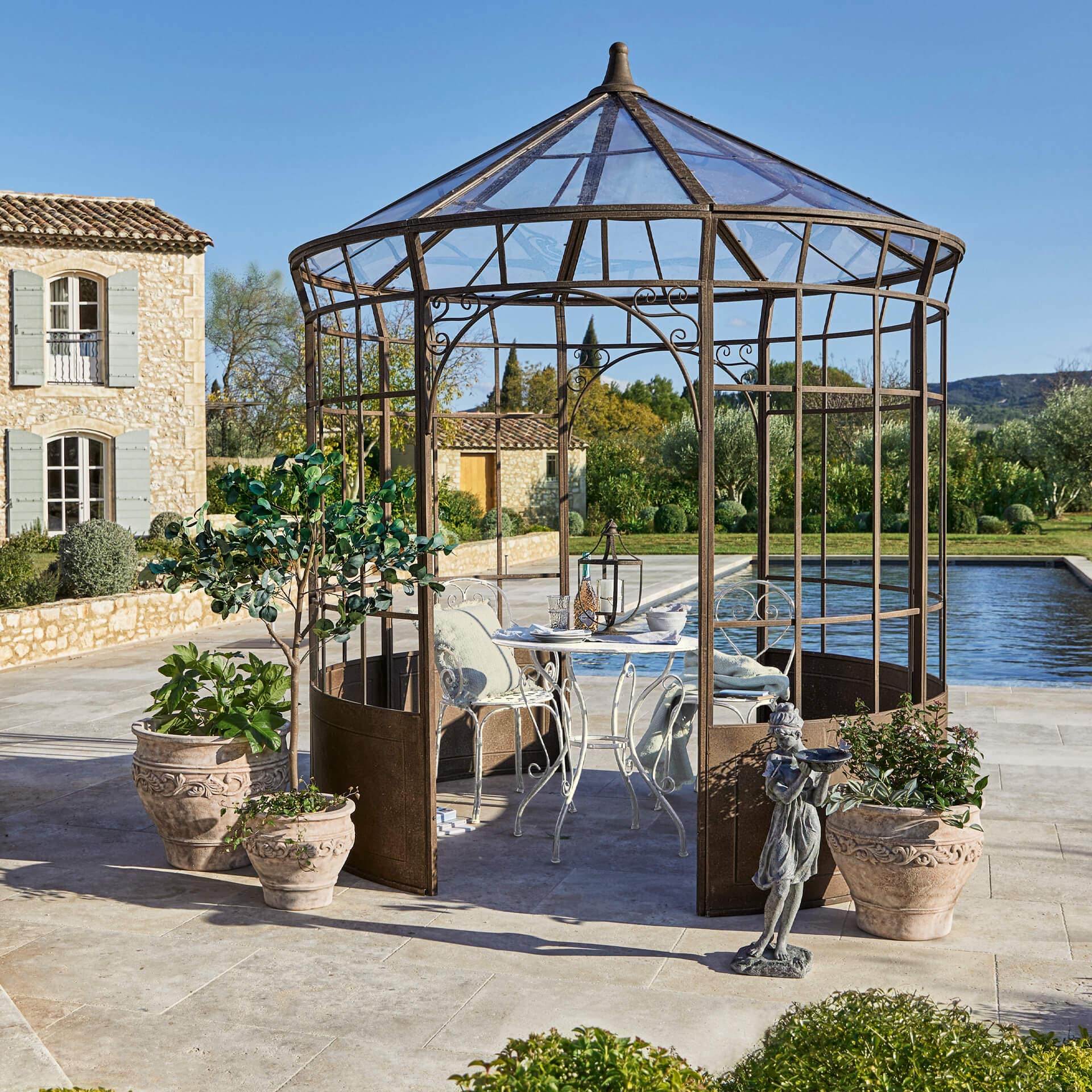 Garten mit rostfarbener Metalllaube, weissem Tisch und Stühlen, umgeben von Pflanzgefässen