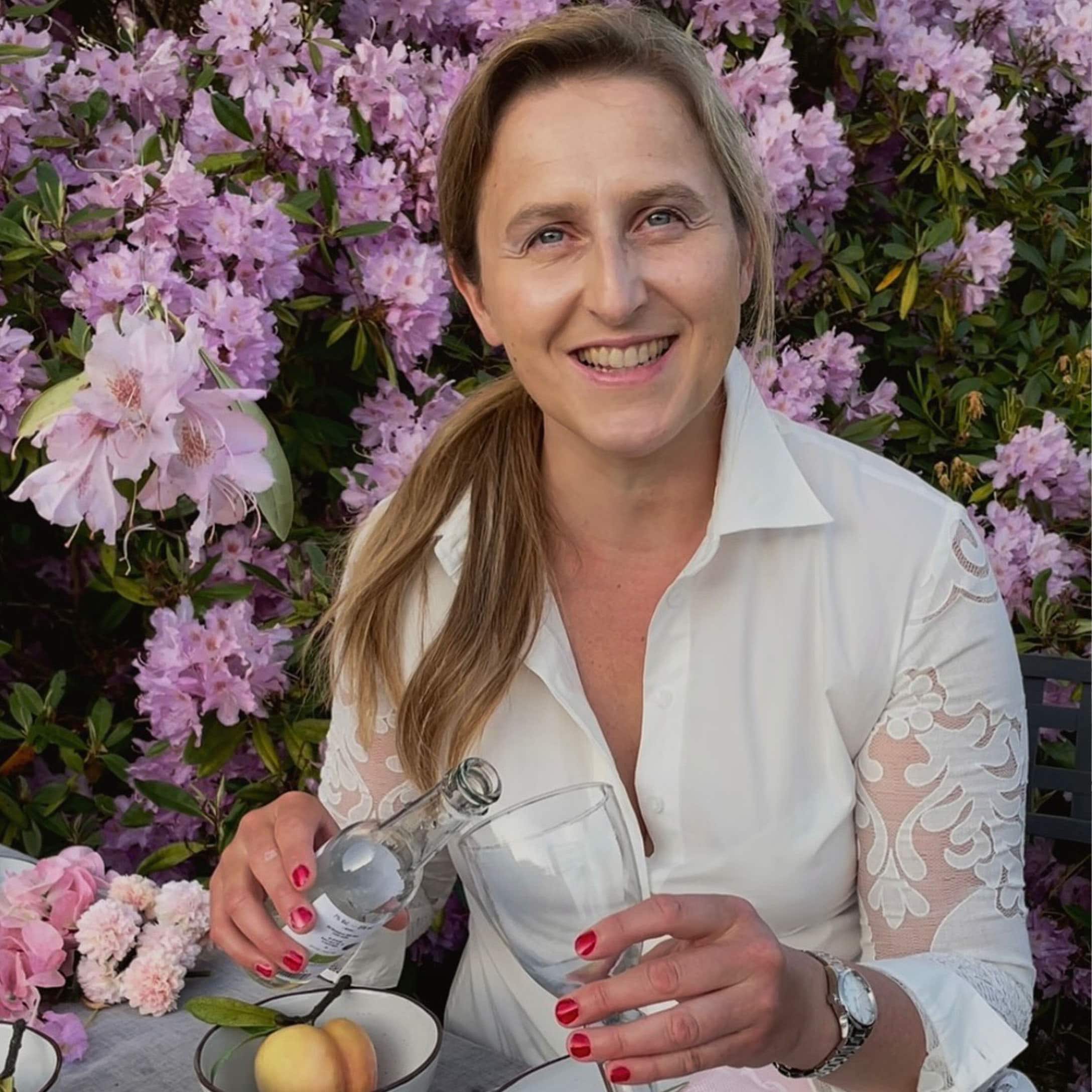 Frau in weisser Bluse mit Spitzenärmeln gießt Getränk in Glas vor rosa Blüten ein.