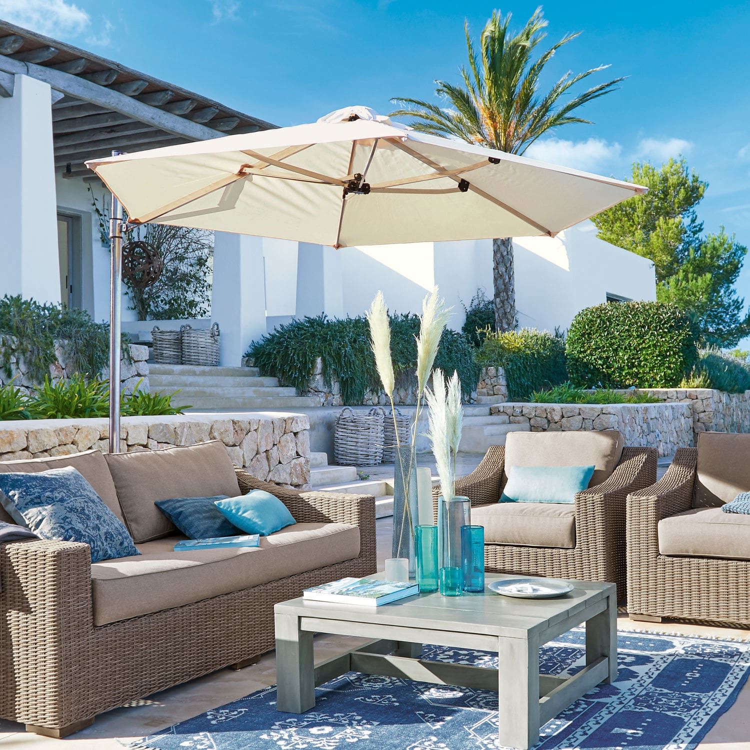 Gartenmöbel-Set aus beigem Polyrattan mit cremefarbenem Sonnenschirm auf Terrasse mit blauem Teppich