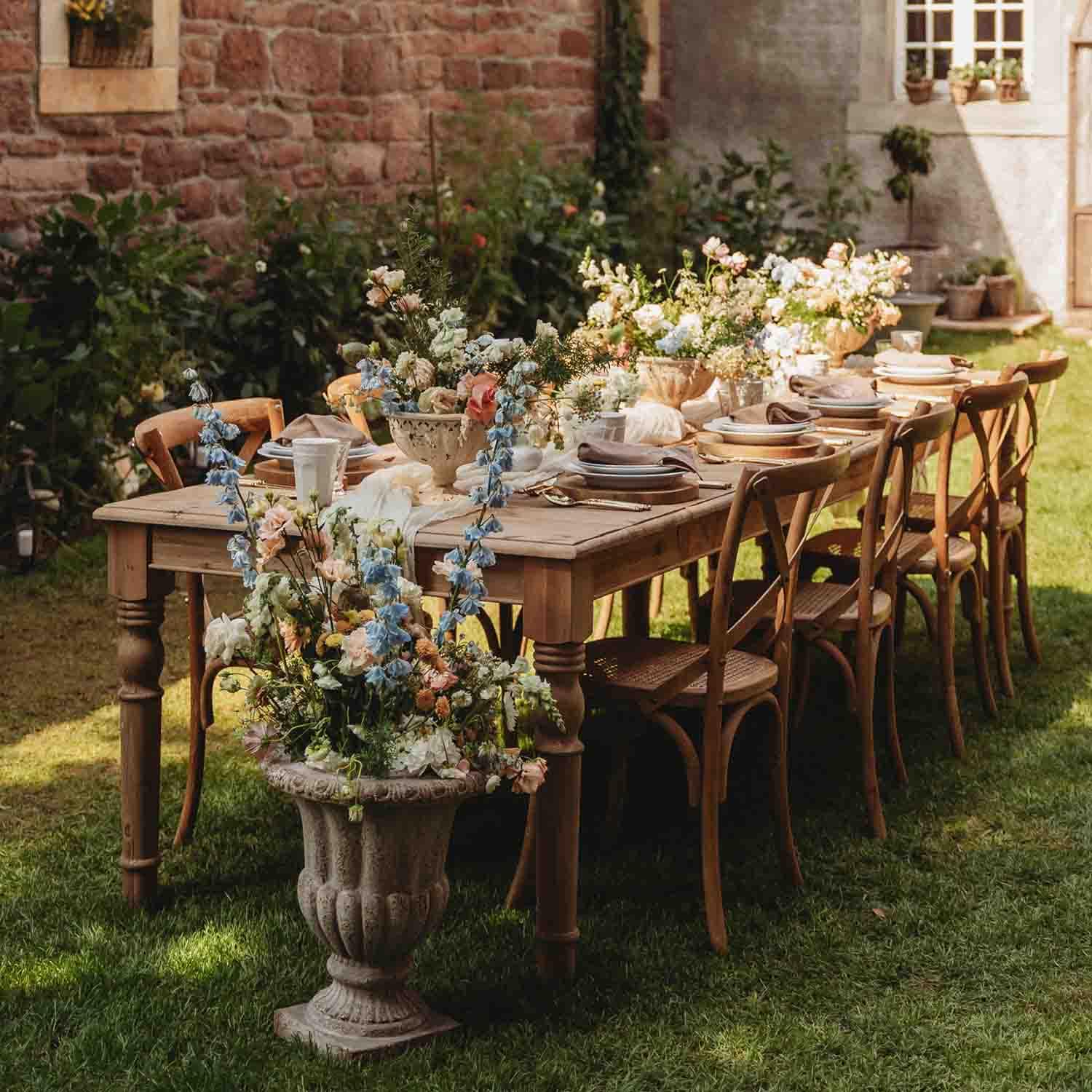 Holztisch im Garten mit Blumendekoration und gedecktem Geschirr für eine Outdoor-Feier