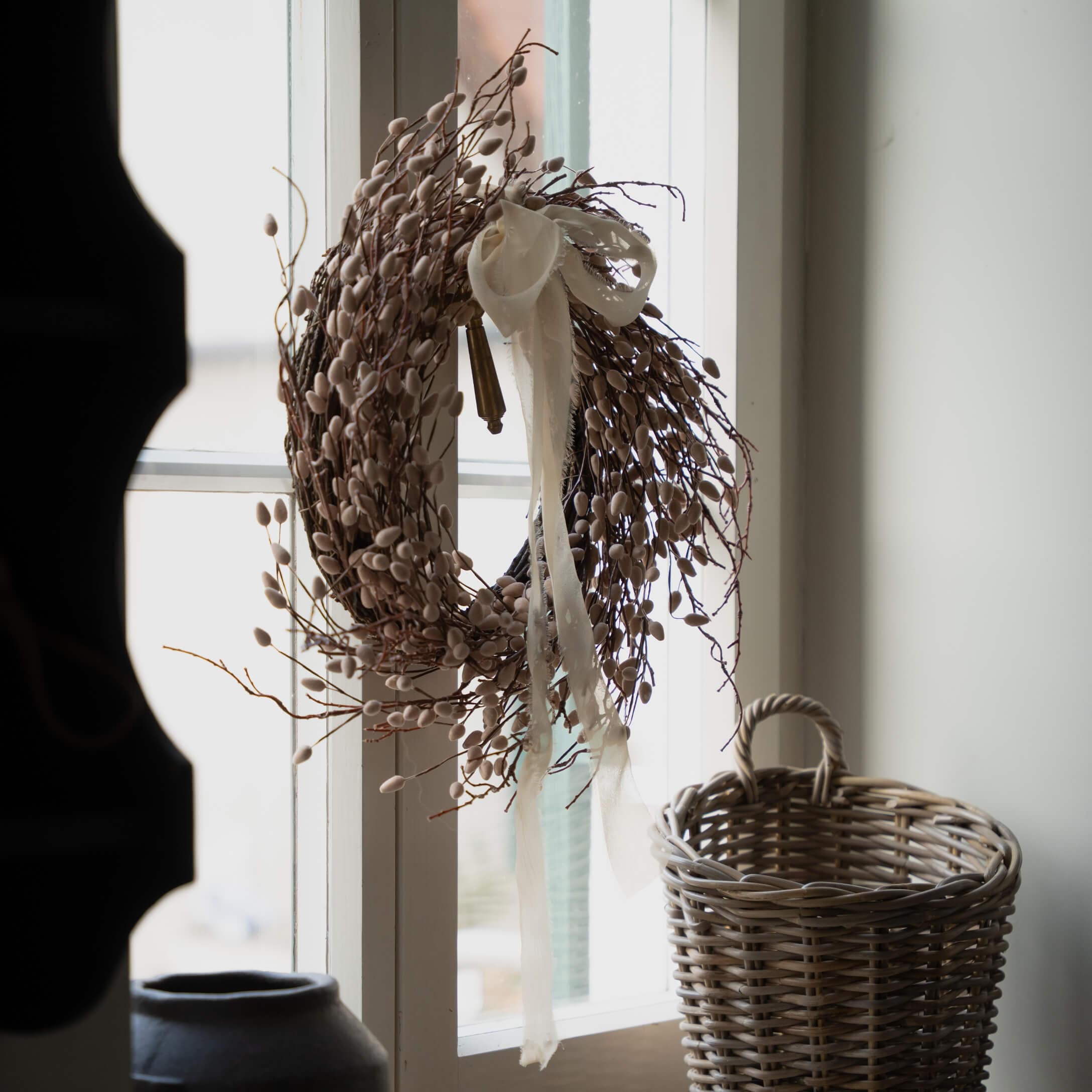 Dekorativer Kranz aus Zweigen und Knospen in Beige mit weissem Band am Fenster, daneben ein Weidenkorb
