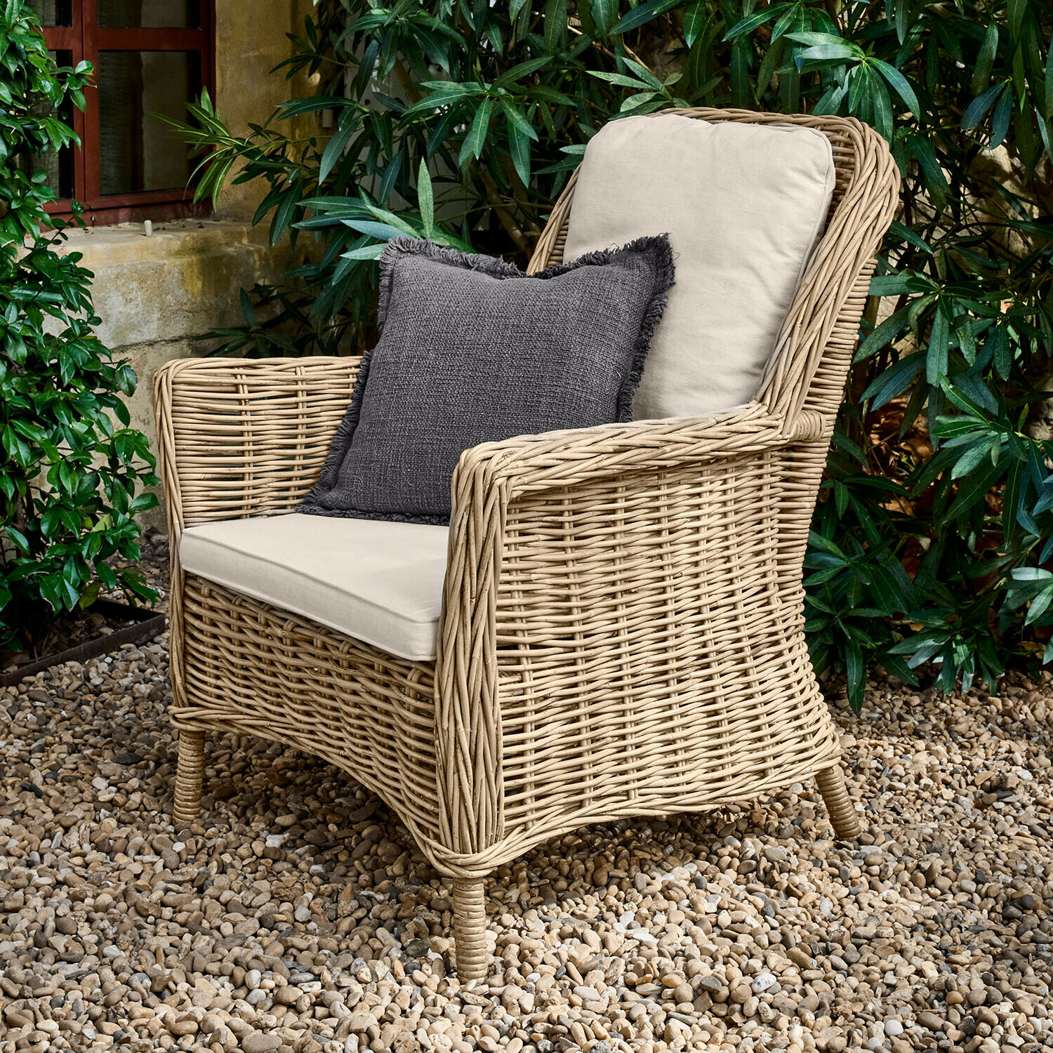 Sessel Travenne Rattansessel mit cremefarbenen Polstern und dunkelgrauem Dekokissen auf Kiesboden im Garten