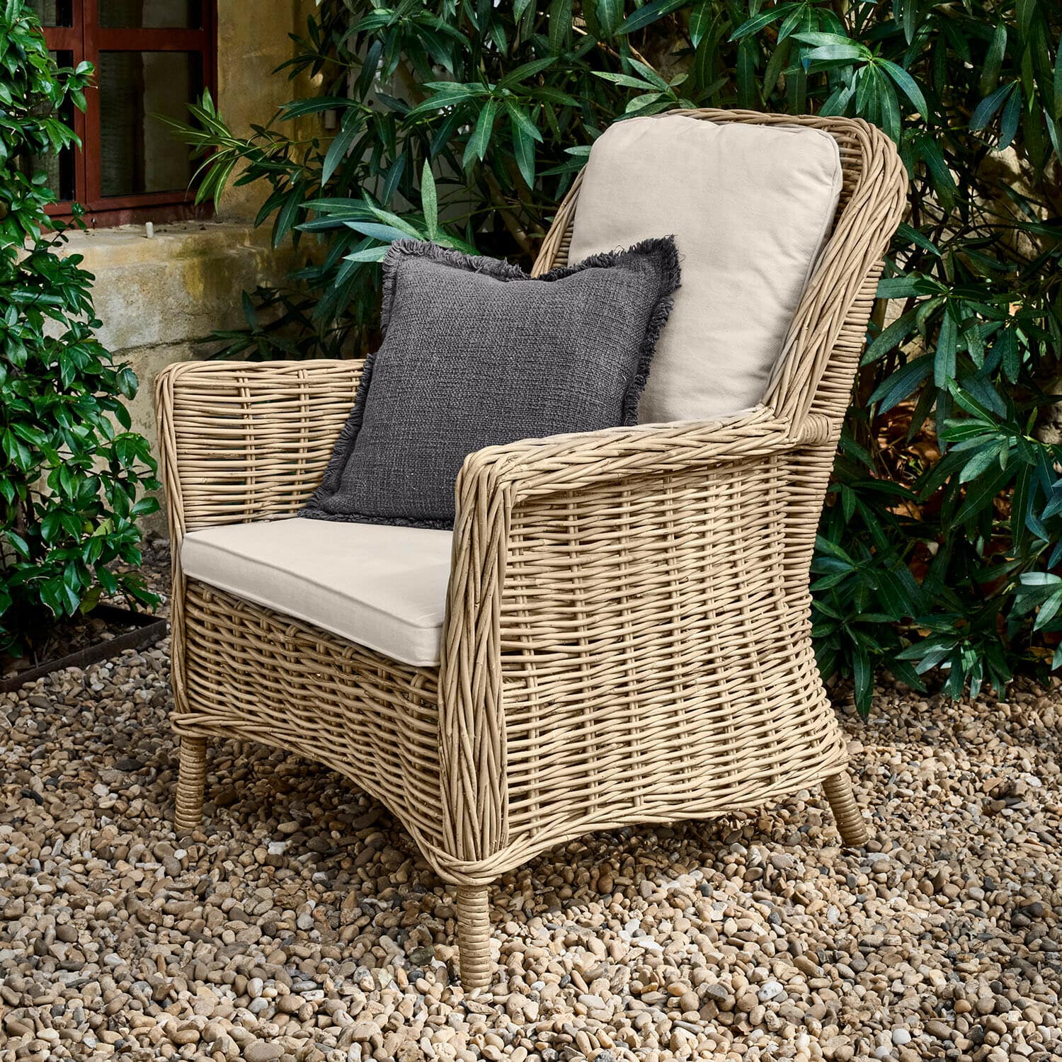 Sessel Travenne Rattansessel mit cremefarbenen Polstern und dunkelgrauem Dekokissen auf Kiesboden im Garten