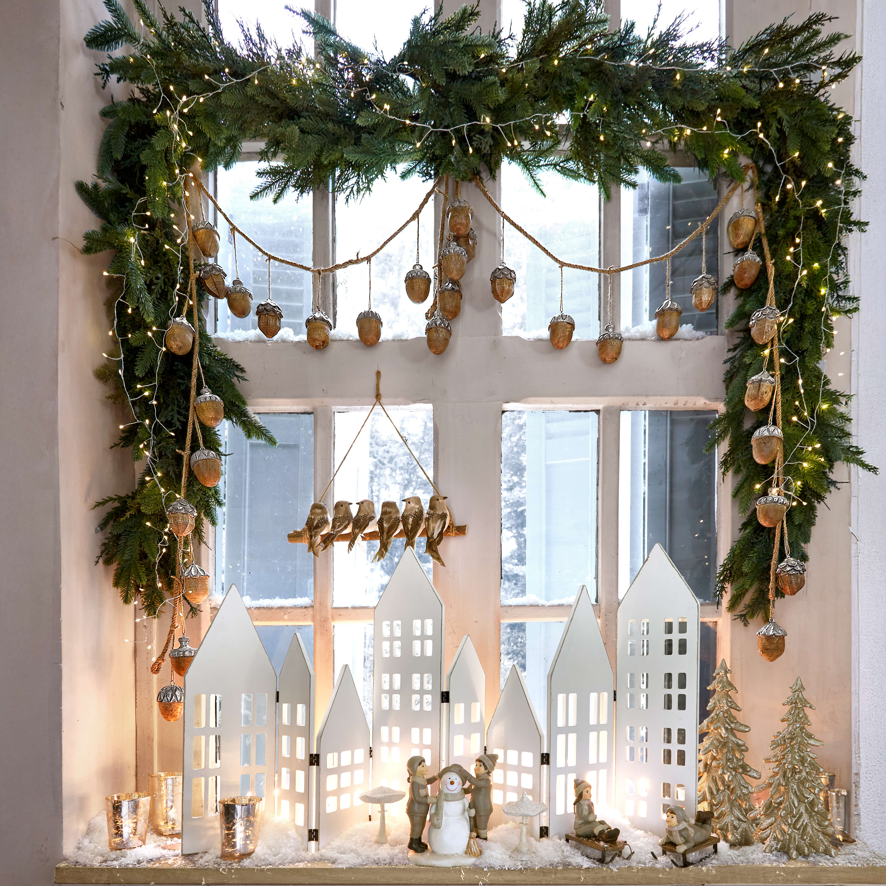 Weihnachtsdekoration mit weissen Häusern, Kinderfiguren und Schneemann auf Fensterbank mit grüner Girlande und Lichterkette