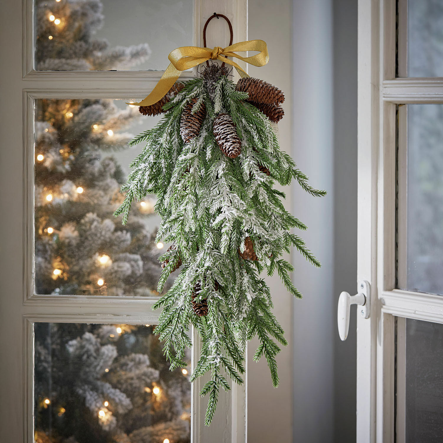Weihnachtliche Hängedekoration aus grünen Zweigen mit Schnee und Zapfen, goldene Schleife