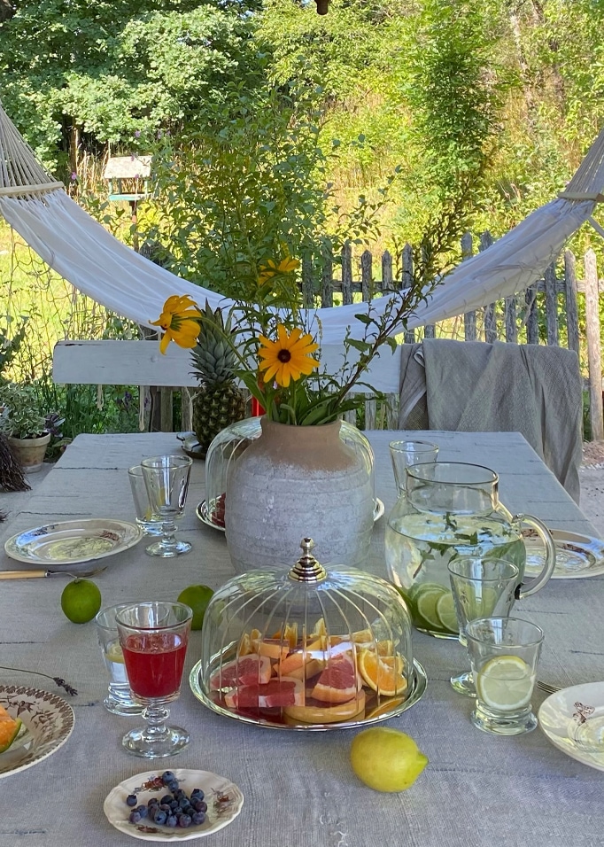 Gartentisch mit grauem Tuch, Vase mit gelben Blumen und Glasglocke mit frischen Früchten.