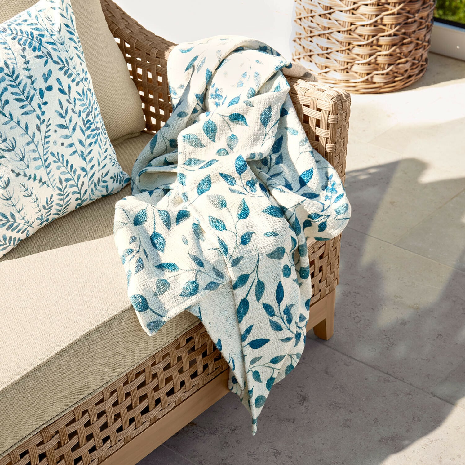 Plaid Ocevara Beiges Sofa mit geflochtenem Rücken und cremefarbiger Decke mit blauem Blattmuster auf Terrasse