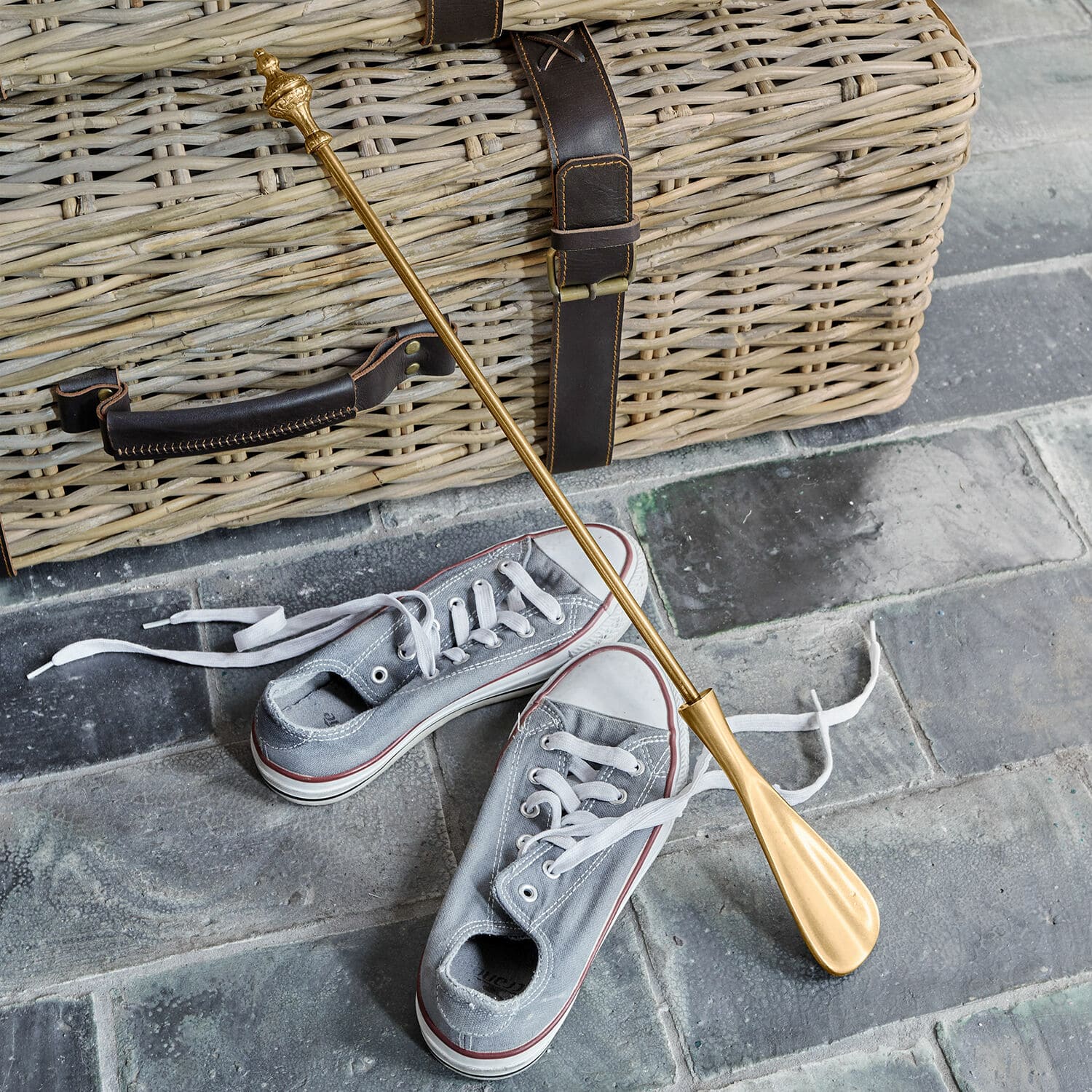 Schuhlöffel Orveline Goldfarbener Metall-Schuhlöffel vor grauen Turnschuhen und einem Korb aus geflochtenem Holz