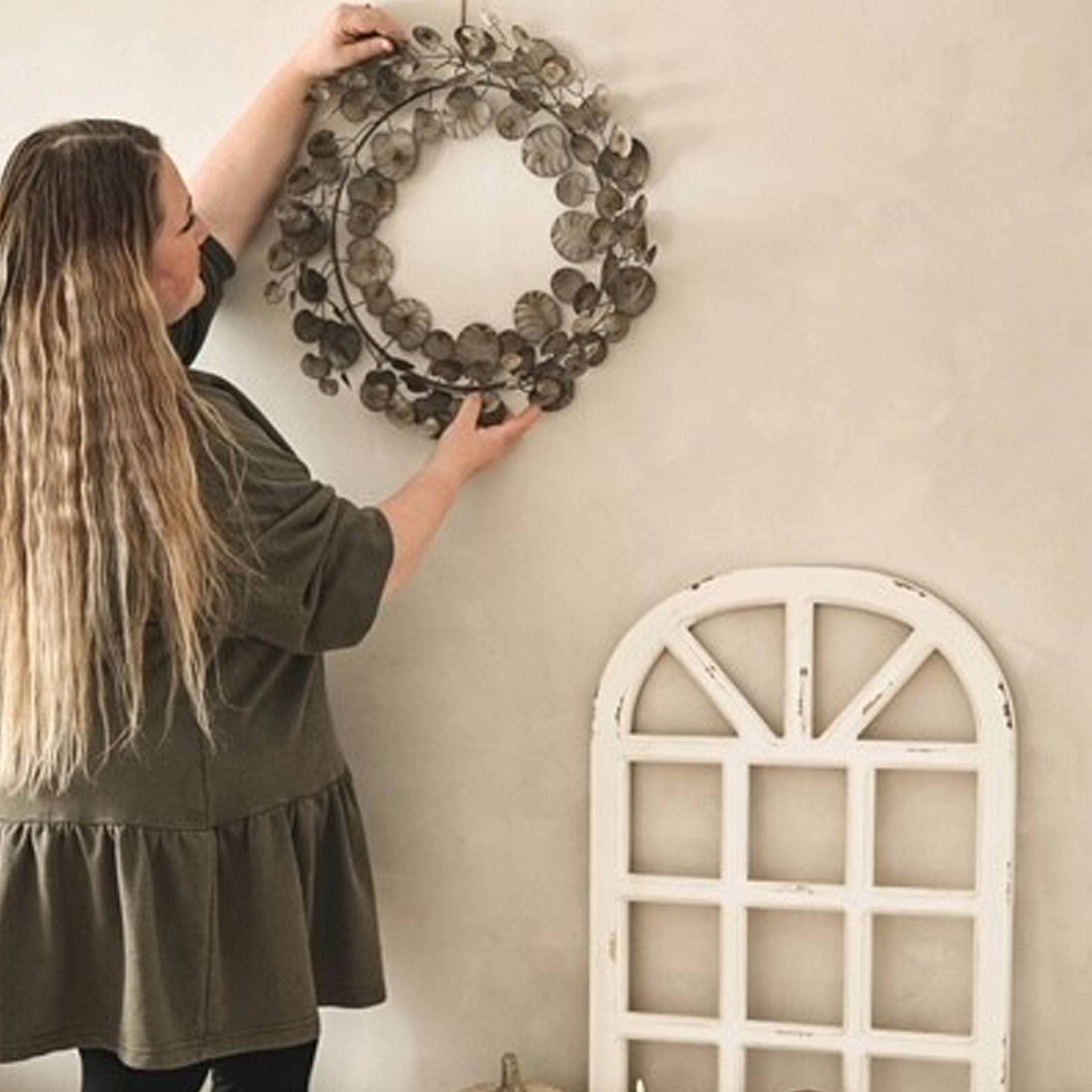 Frau hängt braunen Metall-Dekokranz an helle Wand im rustikalen Stil auf.