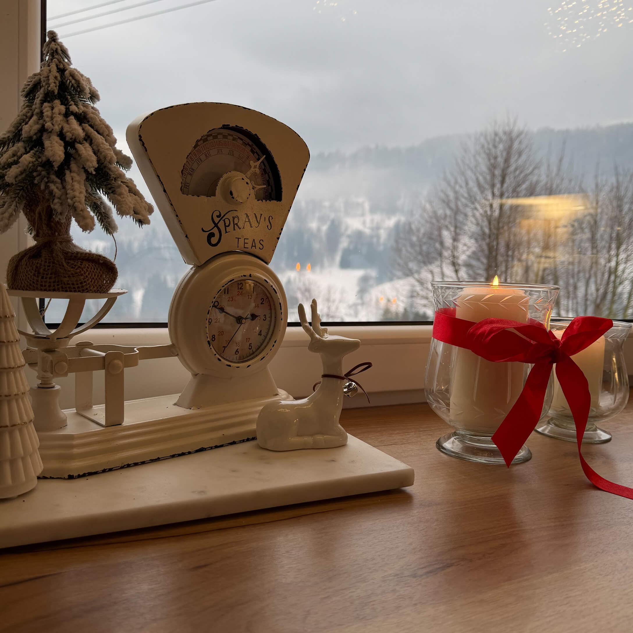 Weisse Metall-Küchenwaage mit Schriftzug Spray's Teas, Weihnachtsdekoration und Kerze mit roter Schleife auf Fensterbank