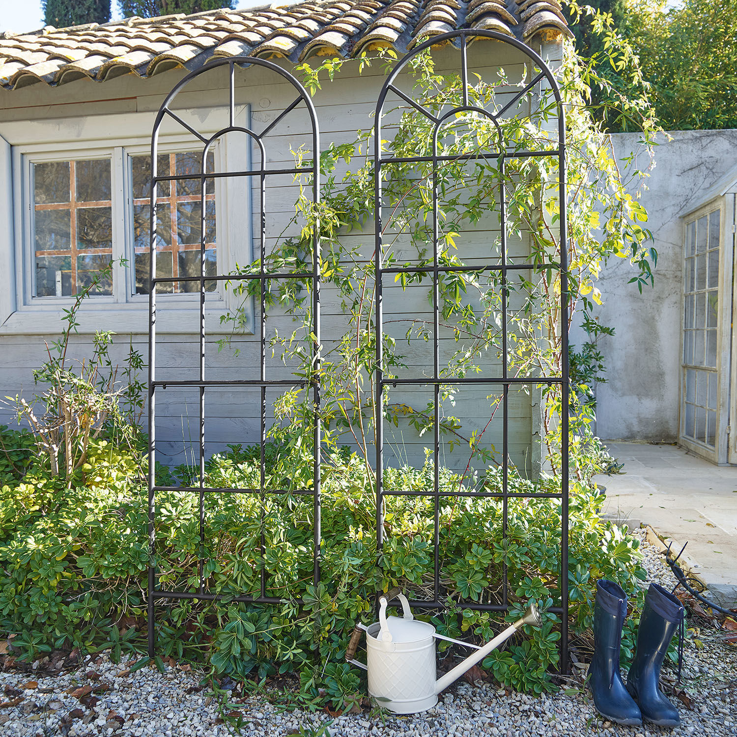 Rankgitter 2er Set Ealdred Schwarzes Metall-Gartenrankgitter mit Bogenmuster vor grünem Bewuchs und Haus, Giesskanne und Gummistiefel