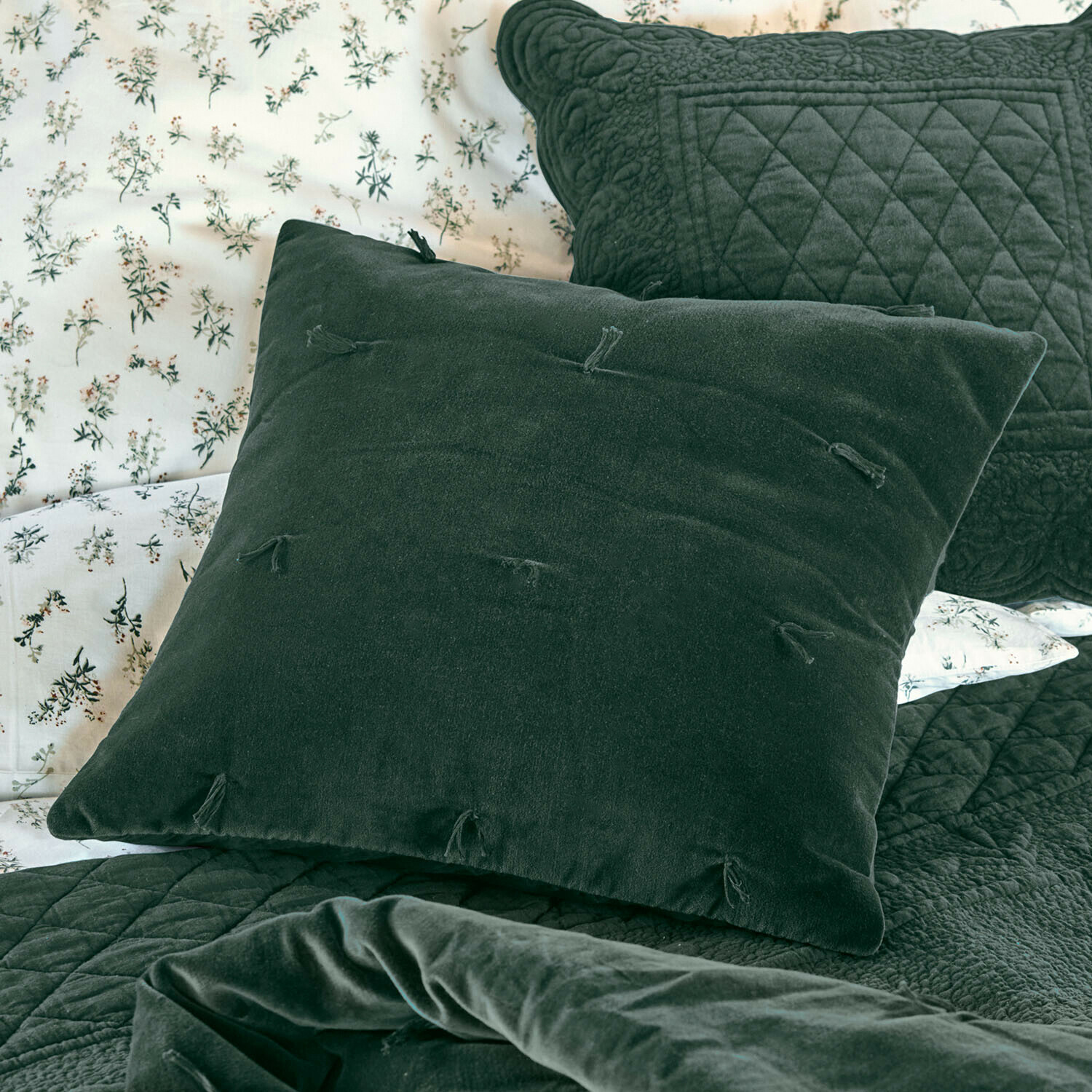 Grünes Samtkissen mit Quasten auf Bett mit gesteppter Decke und floralem Bettbezug
