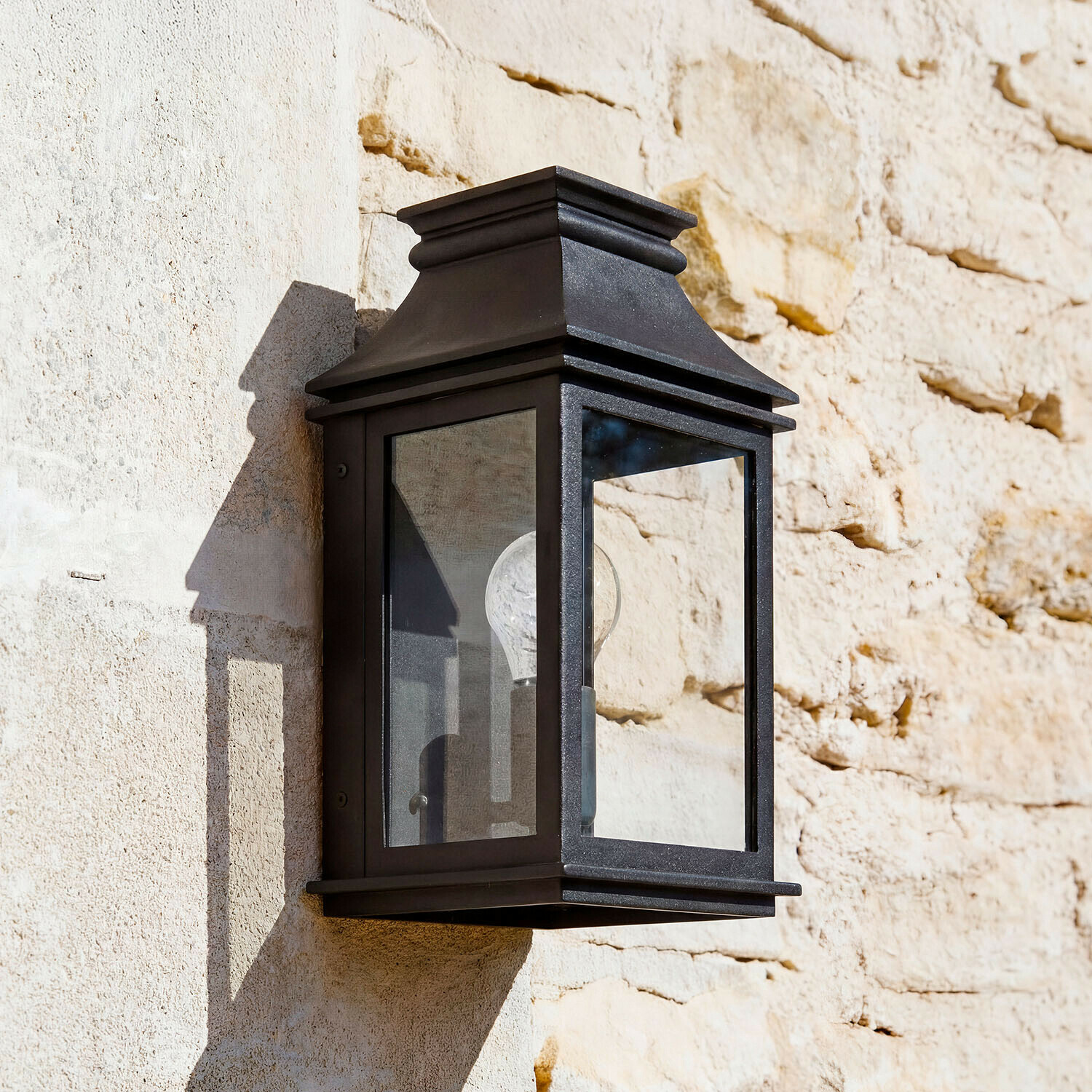 Außenwandlampe Zélivarne Schwarze Metall-Wandleuchte mit klarem Glas an heller Steinwand, klassische Aussenbeleuchtung