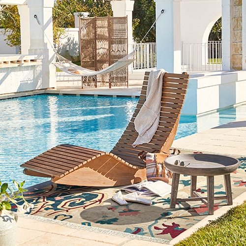 Holzliege auf der Terrasse beim Pool mit buntem Teppich und kleinem Beistelltisch aus Holz