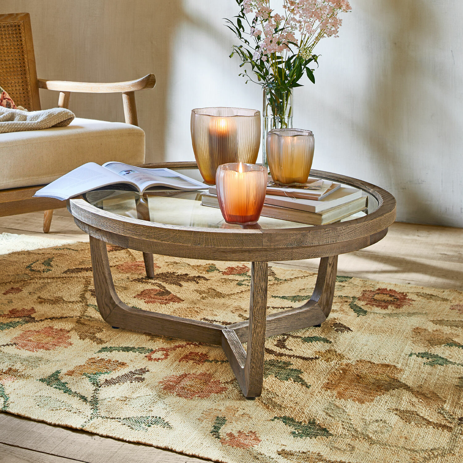 Couchtisch Gradn Enclos Runder Couchtisch aus Holz und Glas auf floralem Teppich, dekoriert mit Kerzen und Büchern