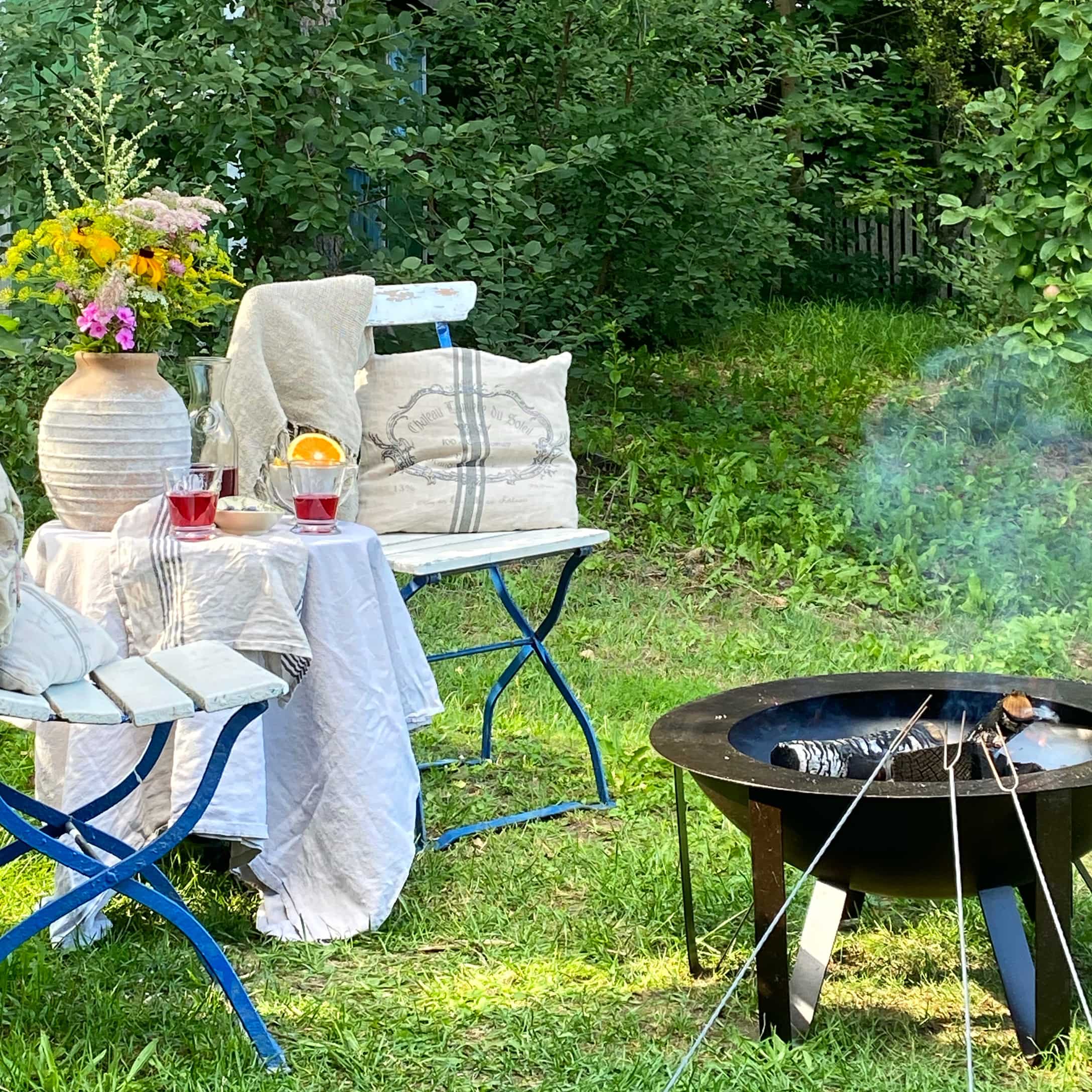 Garten mit blauen Metallstühlen, weiss gedecktem Tisch und schwarzem Feuerkorb mit rauchendem Holz