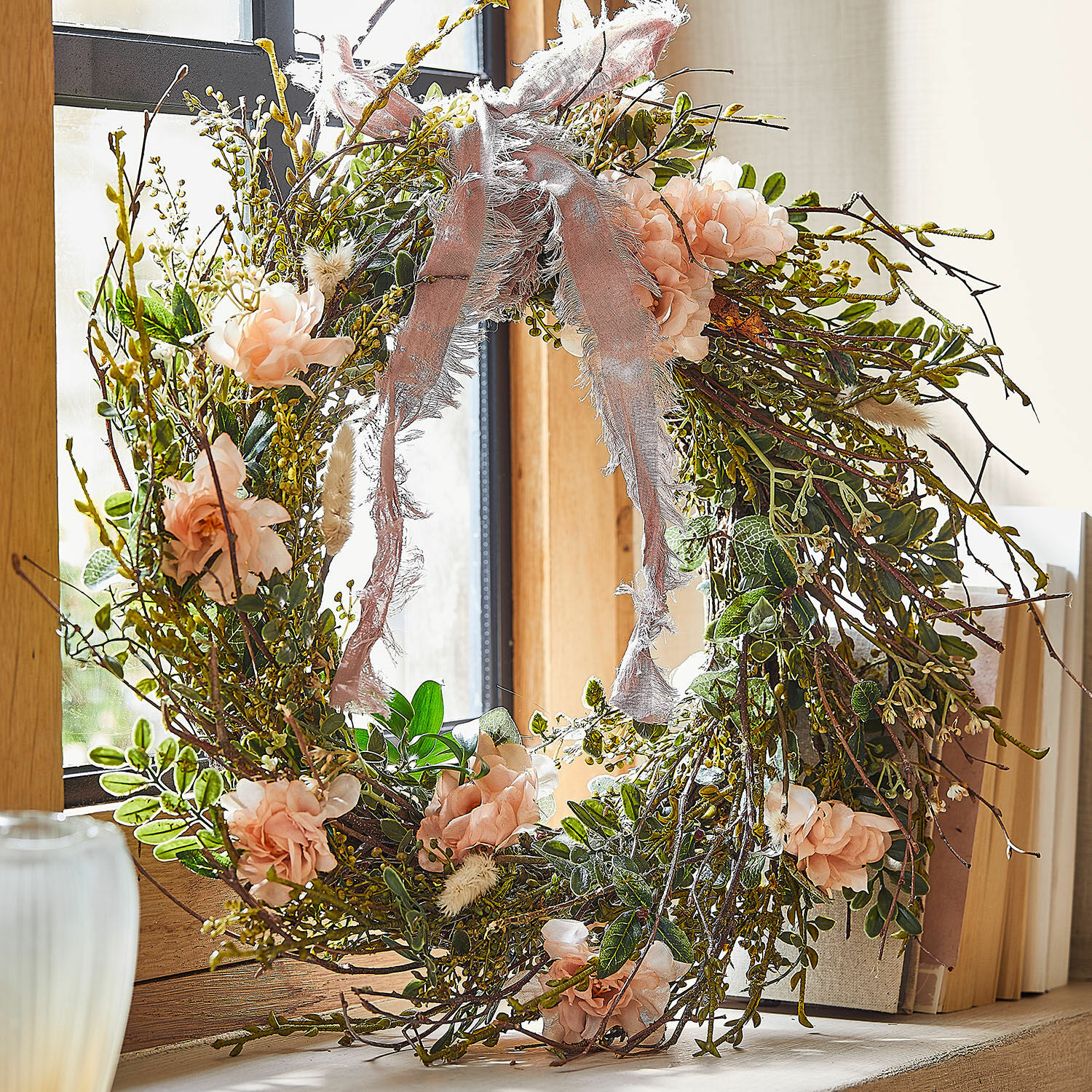 Kranz Chérisse Dekorativer Kranz aus Zweigen, rosa Blüten und grünen Blättern mit rosa Band am Fenster.