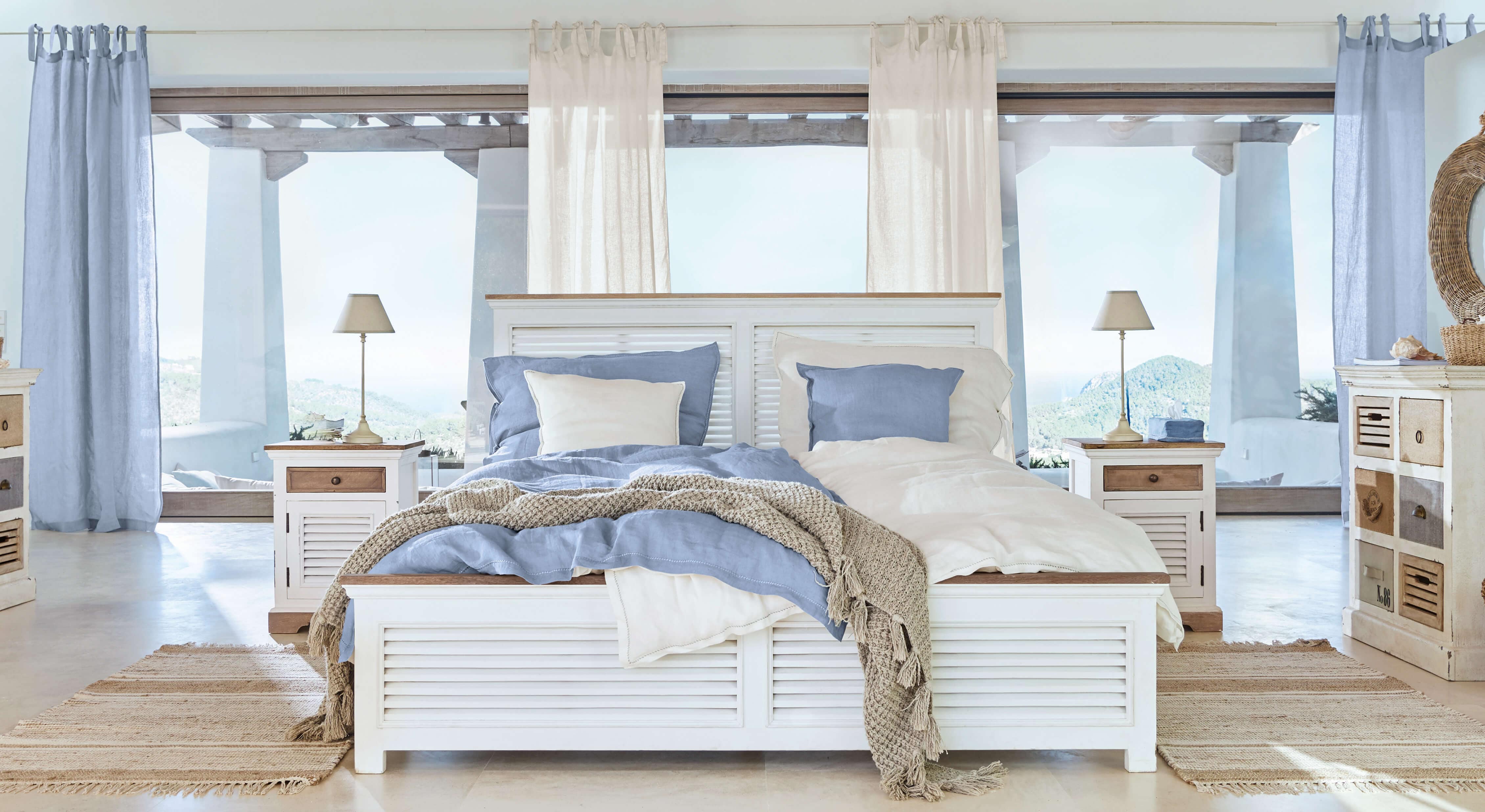 Weisses Holzbett mit blau-beiger Bettwäsche und Naturteppich in hellem Schlafzimmer mit Aussicht
