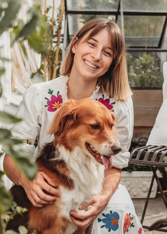 Frau in weissem Kleid mit bunten Stickereien hält braun-weissen Hund in hellem Raum mit Pflanzen.