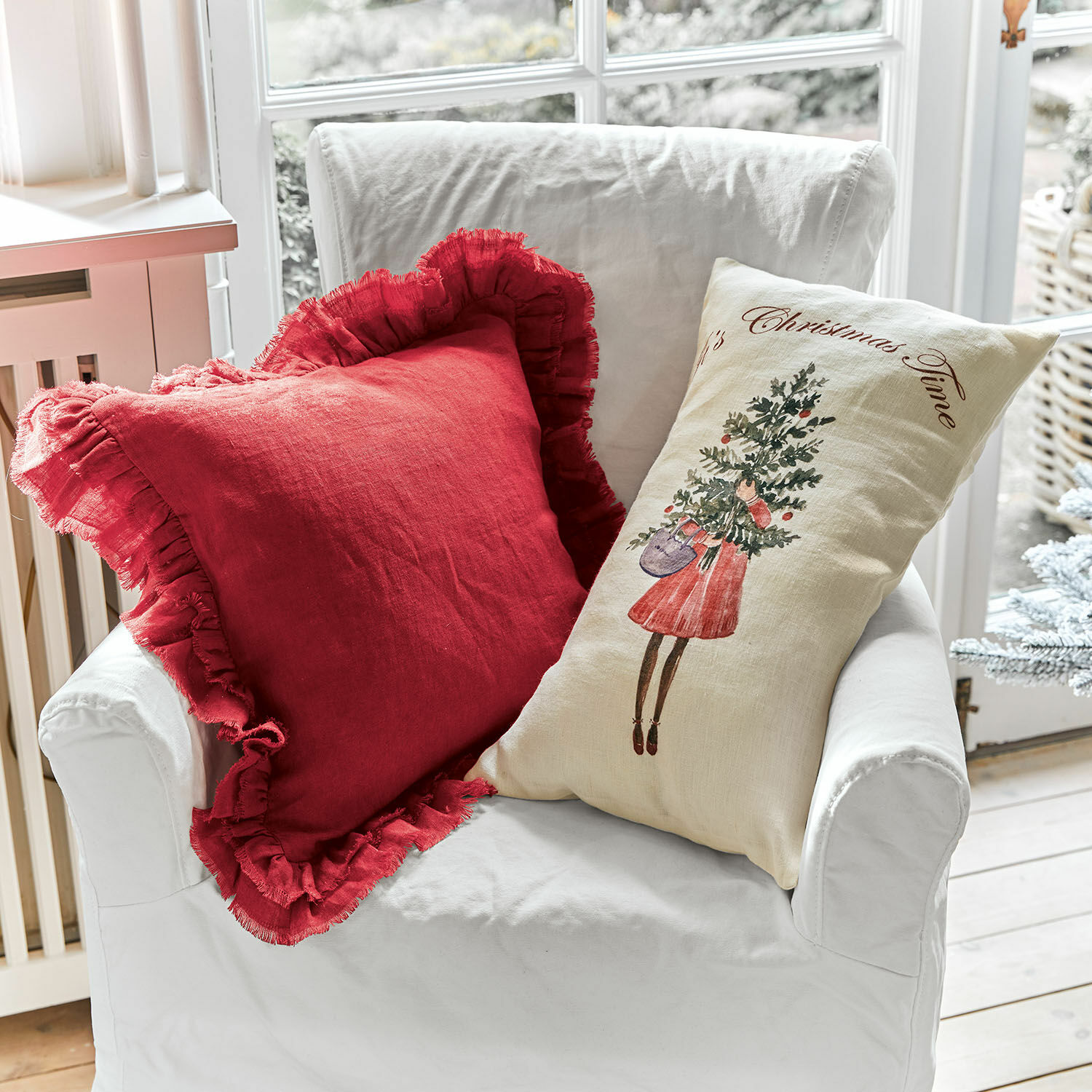 Kissenhülle 2er Set Christmas Time Zwei dekorative Leinenkissen auf weissem Sessel, ein rotes mit Rüschen und ein cremefarbenes mit Weihnachtsmotiv