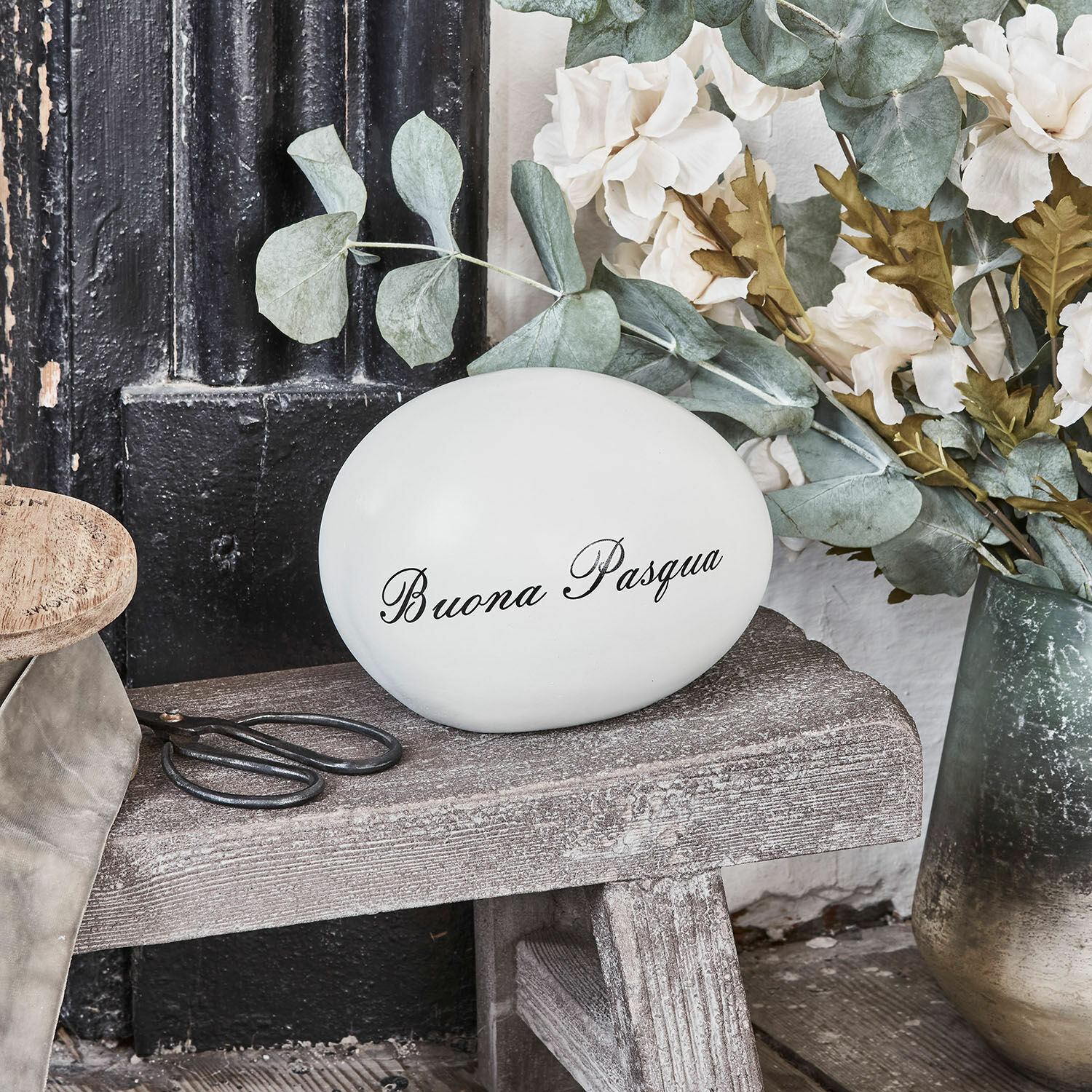 Weiße Keramik-Eierdekoration mit Schriftzug Buona Pasqua auf Holzbank neben Blumenarrangement