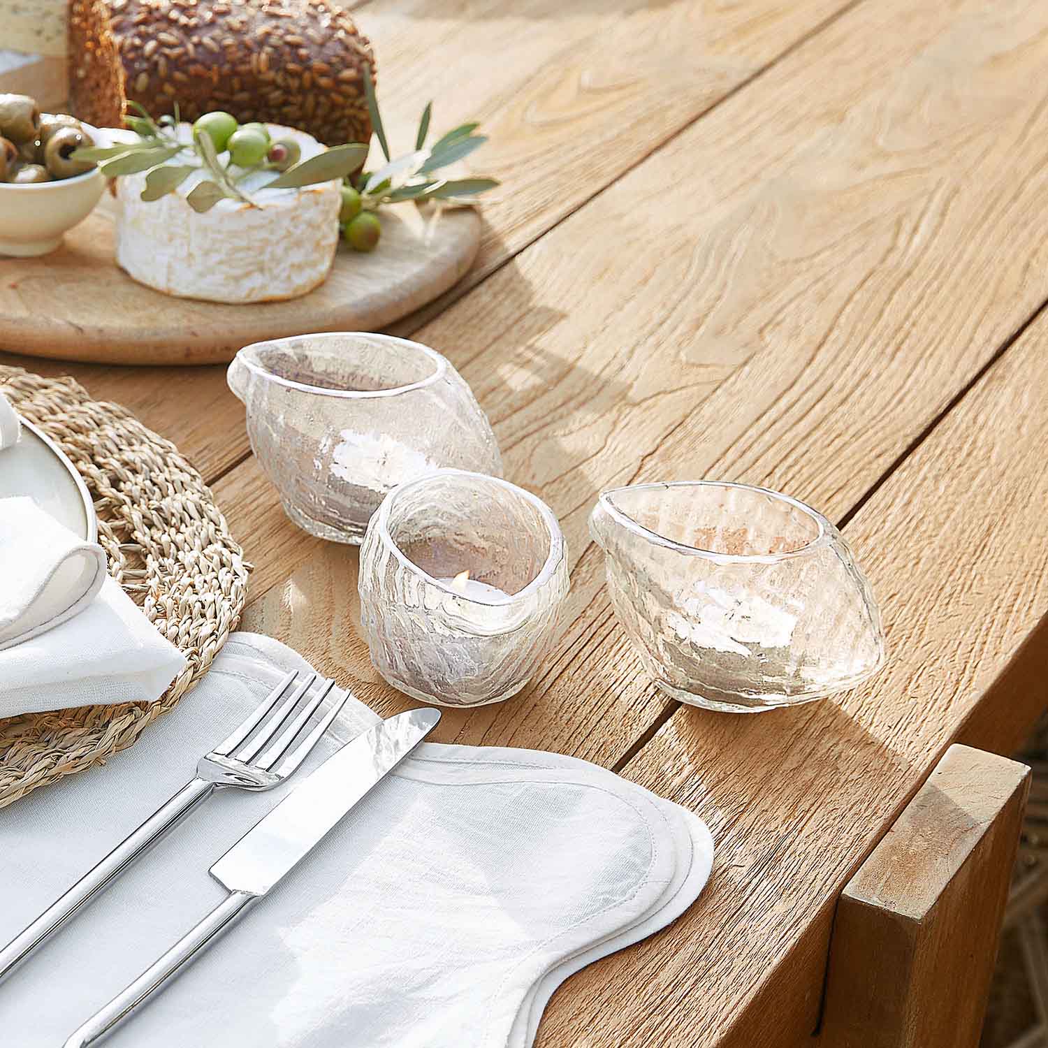 Set mit drei transparenten Glas-Teelichthaltern in unregelmässiger Form auf hellem Holztisch