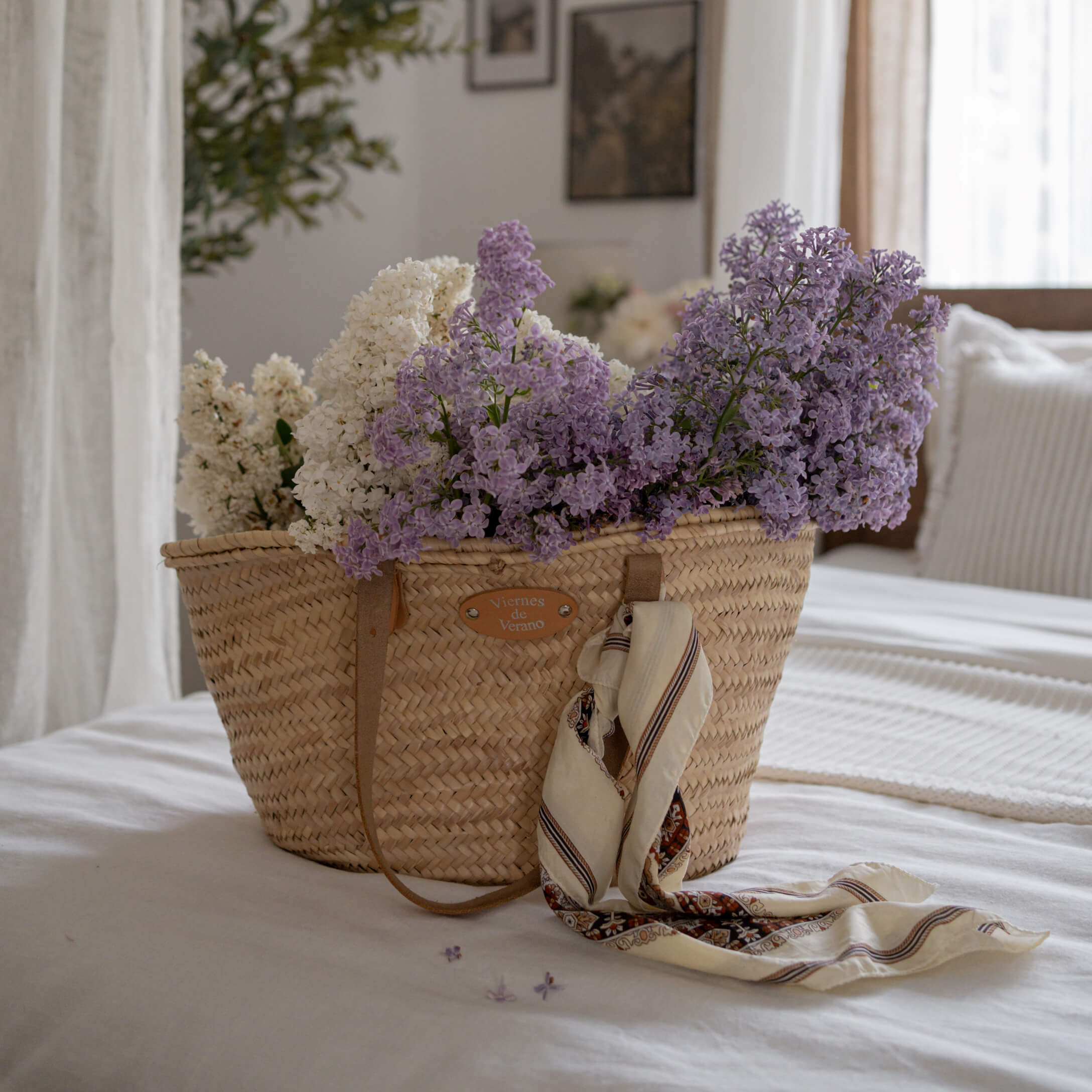 Geflochtener Korb mit weissen und violetten Blumen sowie Tuch auf weissem Bett in hellem Zimmer