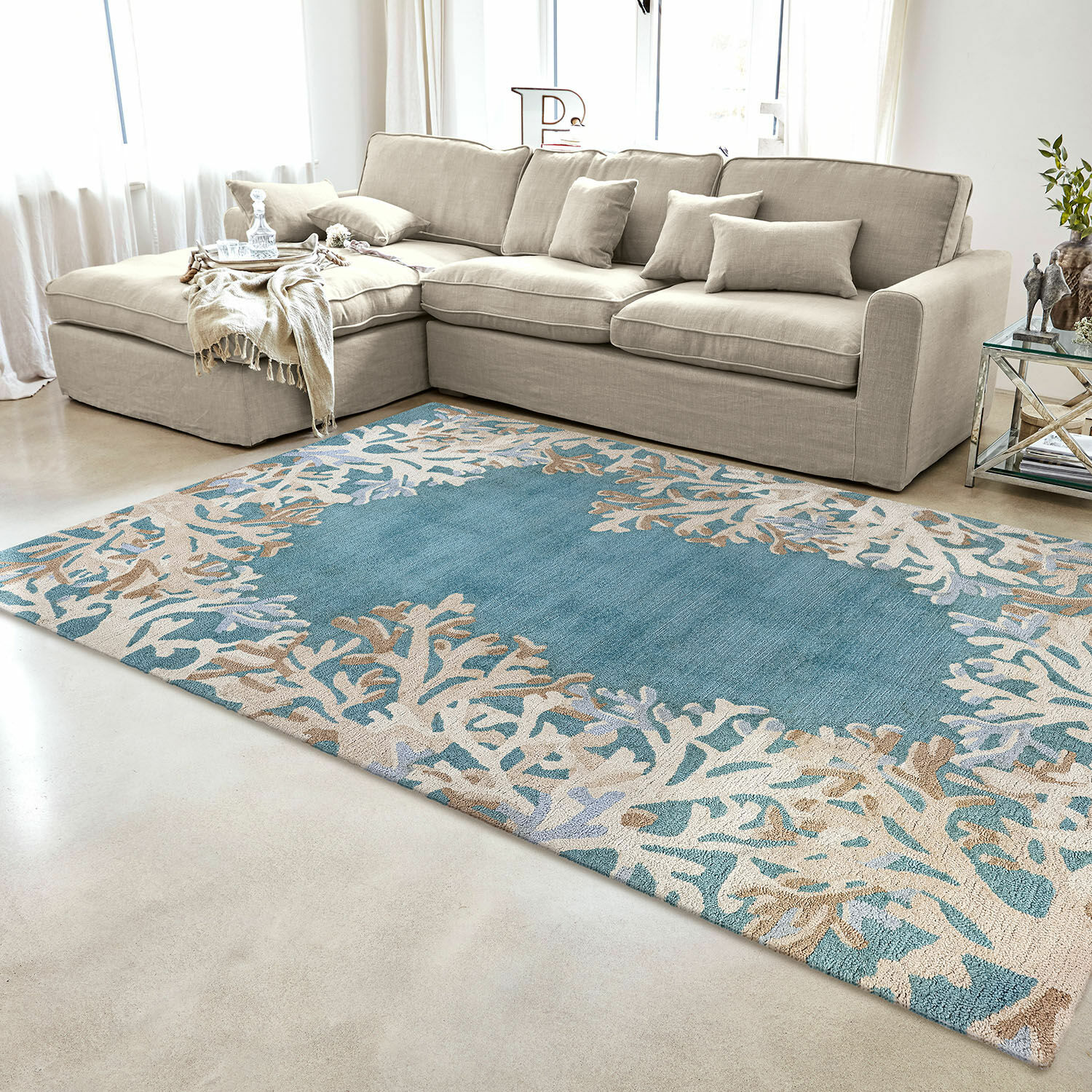 In- & Outdoorteppich Dorlancourt Blau-beiger Teppich mit Korallenmuster im modernen Wohnzimmer mit beigem Sofa und Glastisch.