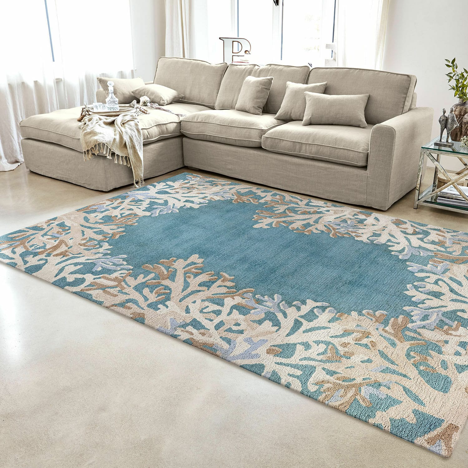 In- & Outdoorteppich Dorlancourt Blau-beiger Teppich mit Korallenmuster im modernen Wohnzimmer mit beigem Sofa und Glastisch.