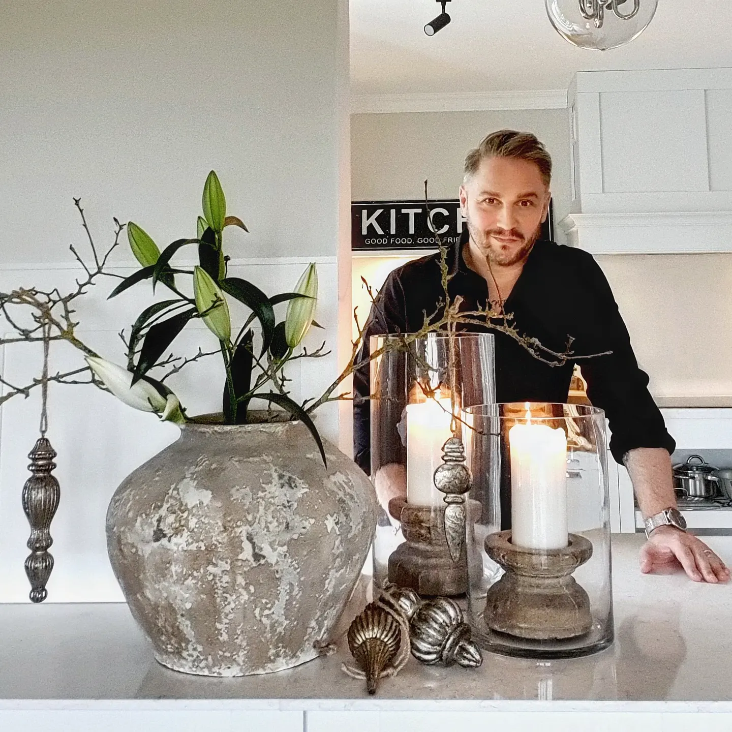 Grosse graue Keramikvase mit grünen Blättern und brennenden Kerzen in Glaswindlichtern auf der Küchenarbeitsplatte