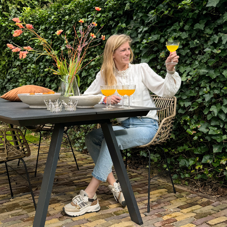 Frau in weisser Bluse und Jeans sitzt draussen an schwarzem Tisch, hält Glas mit orangem Getränk, Vase mit Blumen daneben