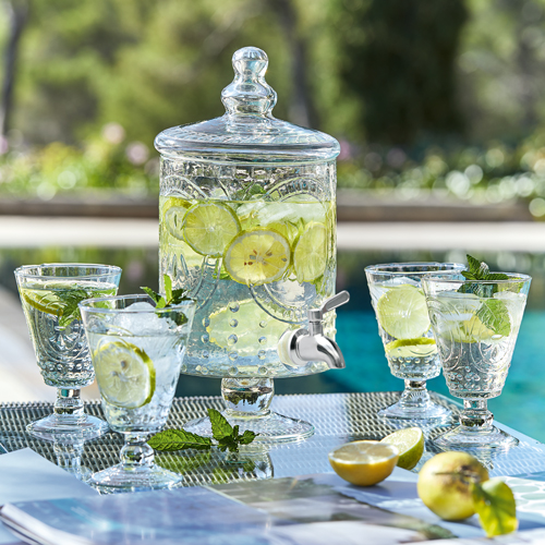 Glasgetränkespender mit Zitronenscheiben und Minze, vier Gläser auf Gartentisch beim Pool