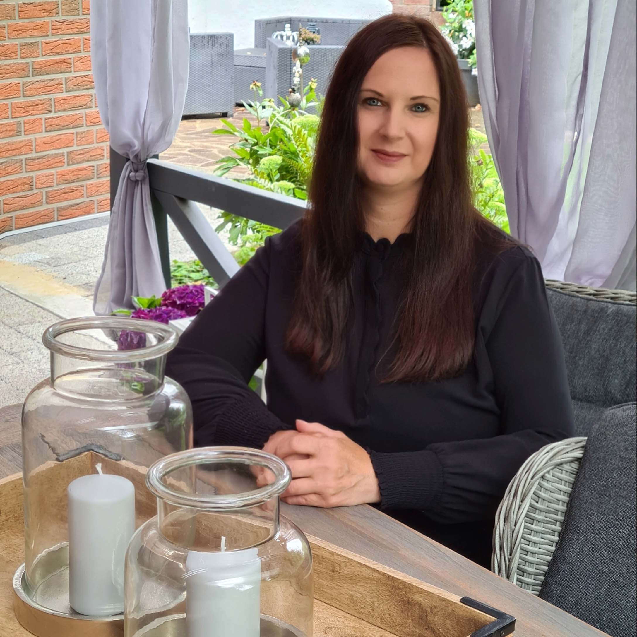 Frau in schwarzer Bluse sitzt an Holztisch mit dekorativen Kerzen in Glaslaternen auf der Terrasse.