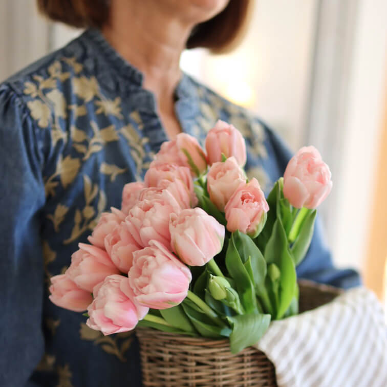 Strauß rosa Tulpen in Weidenkorb gehalten von Frau in blauem Blusenhemd mit Blumenmuster