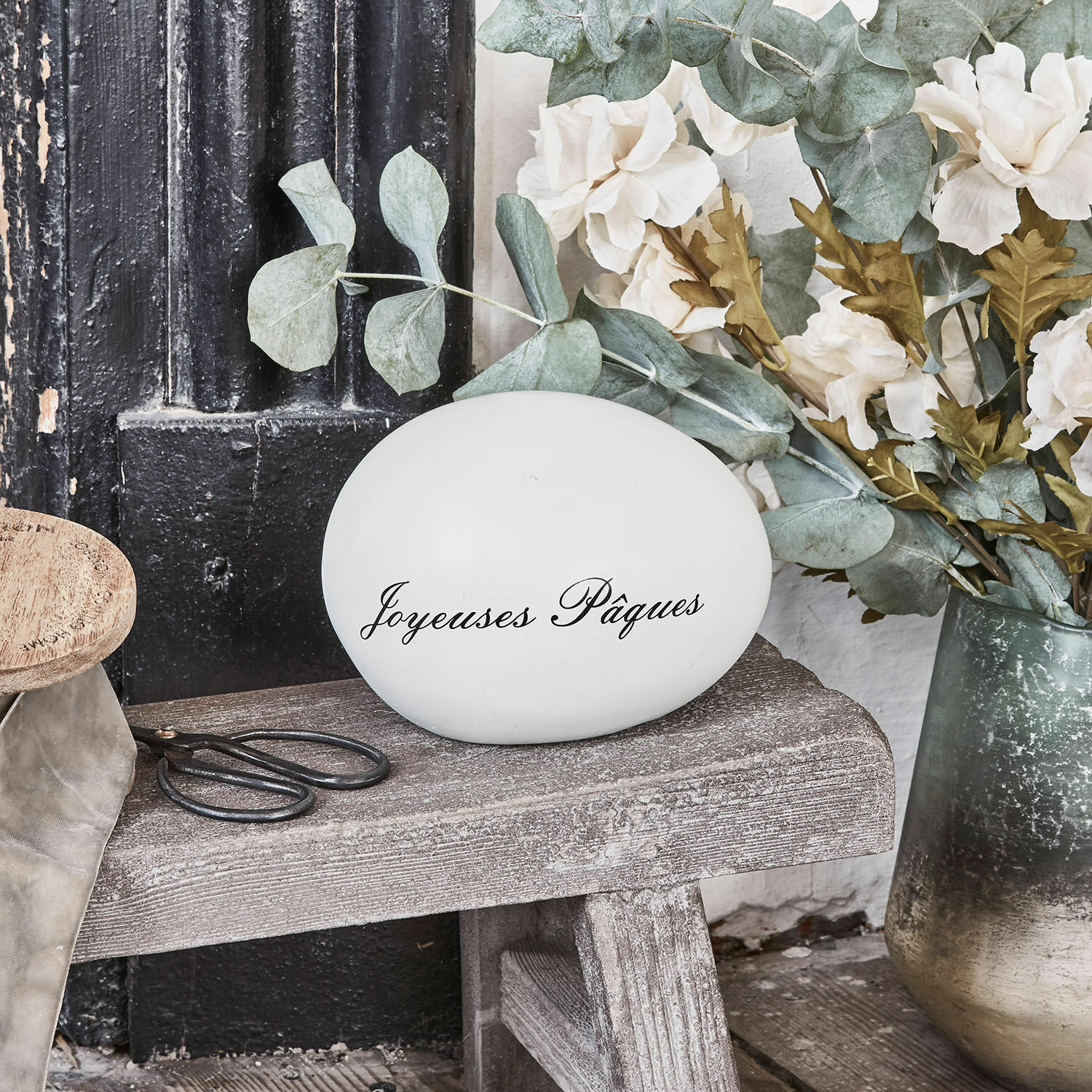 Weisses Dekoei mit Schriftzug Joyeuses Pâques auf Holzbank mit Blumen und Schere