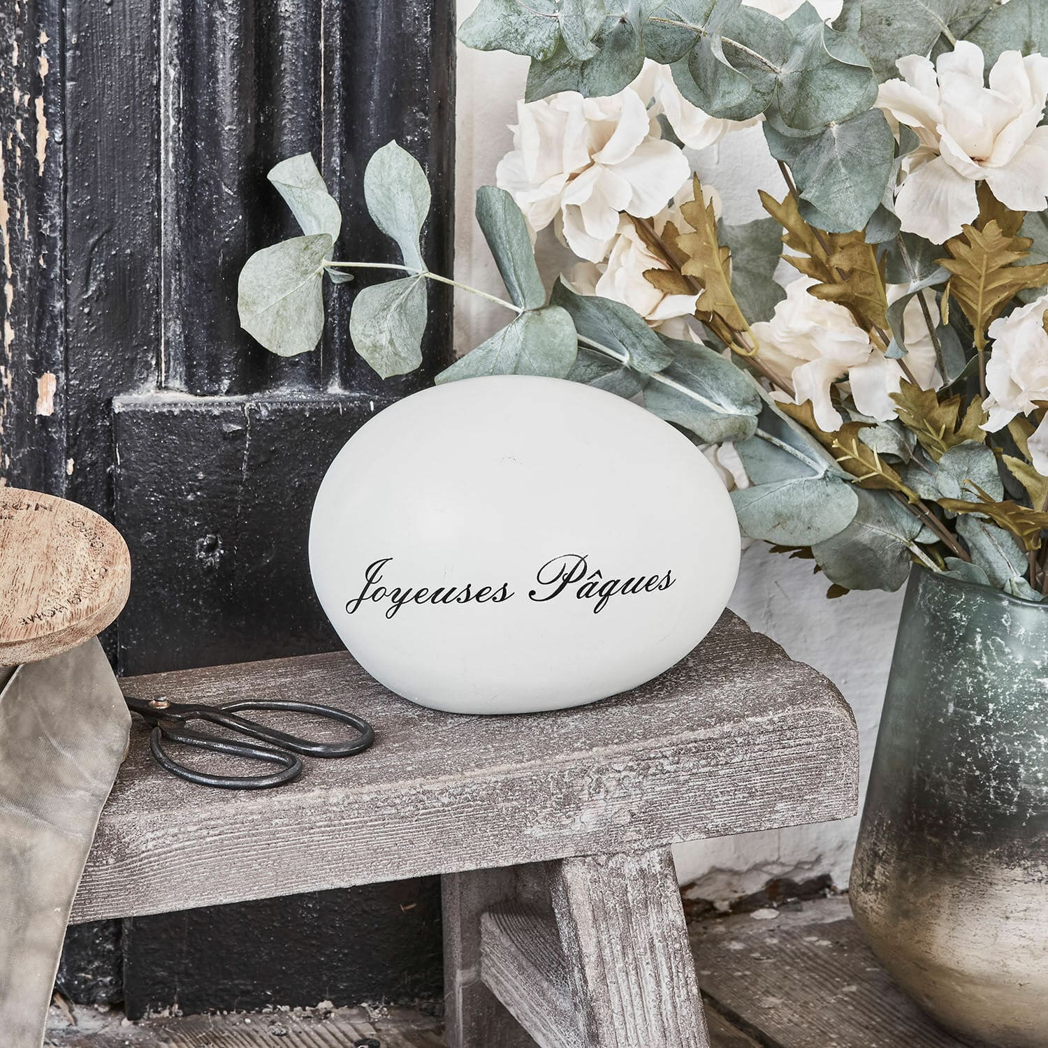 Deko-Ei JOYEUSES PAQUES Weisses Dekoei mit Schriftzug Joyeuses Pâques auf Holzbank mit Blumen und Schere