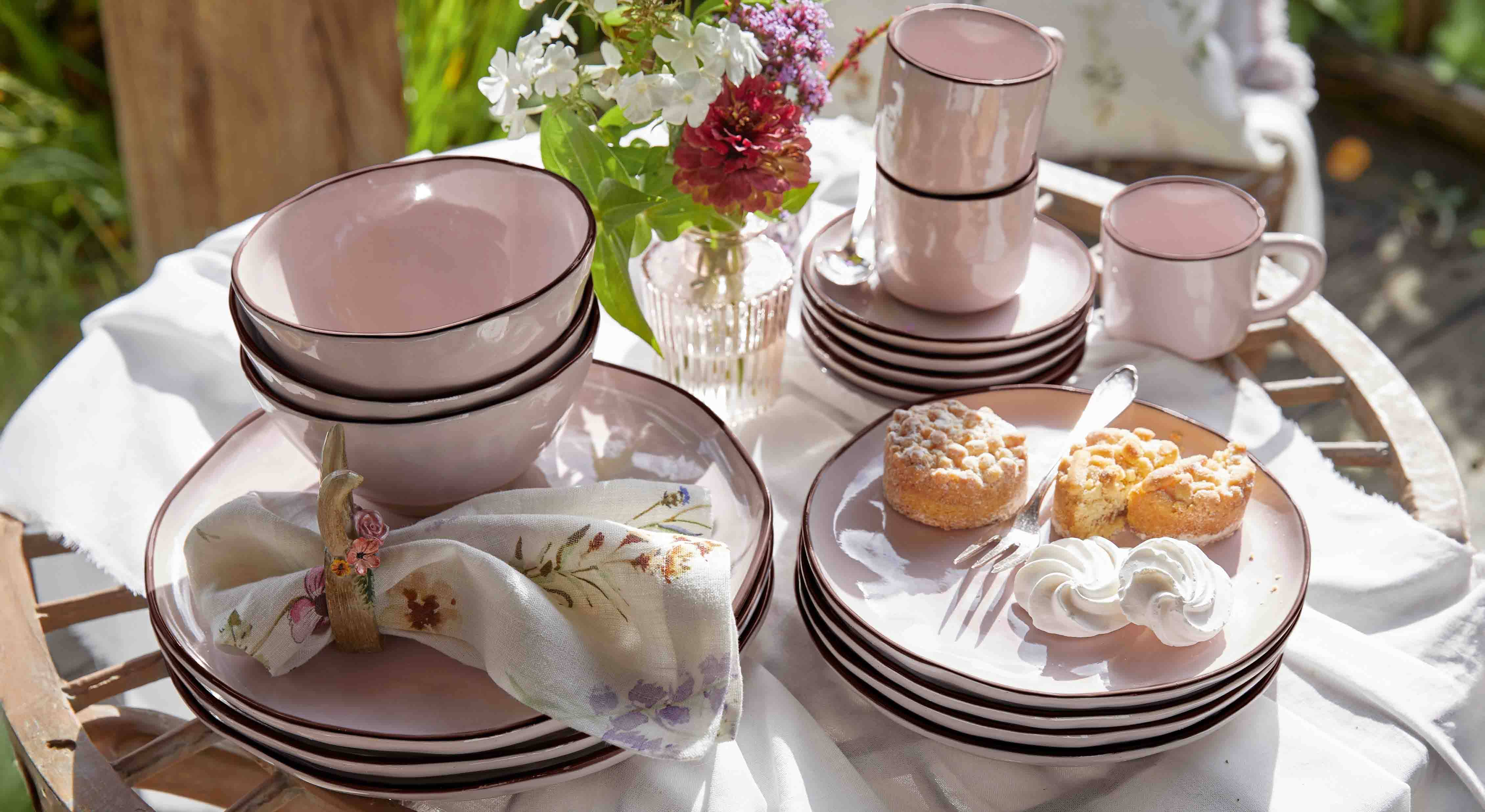Set aus rosa Keramikgeschirr mit Tellern, Schalen und Tassen auf Holztisch mit Kuchen und Blumen