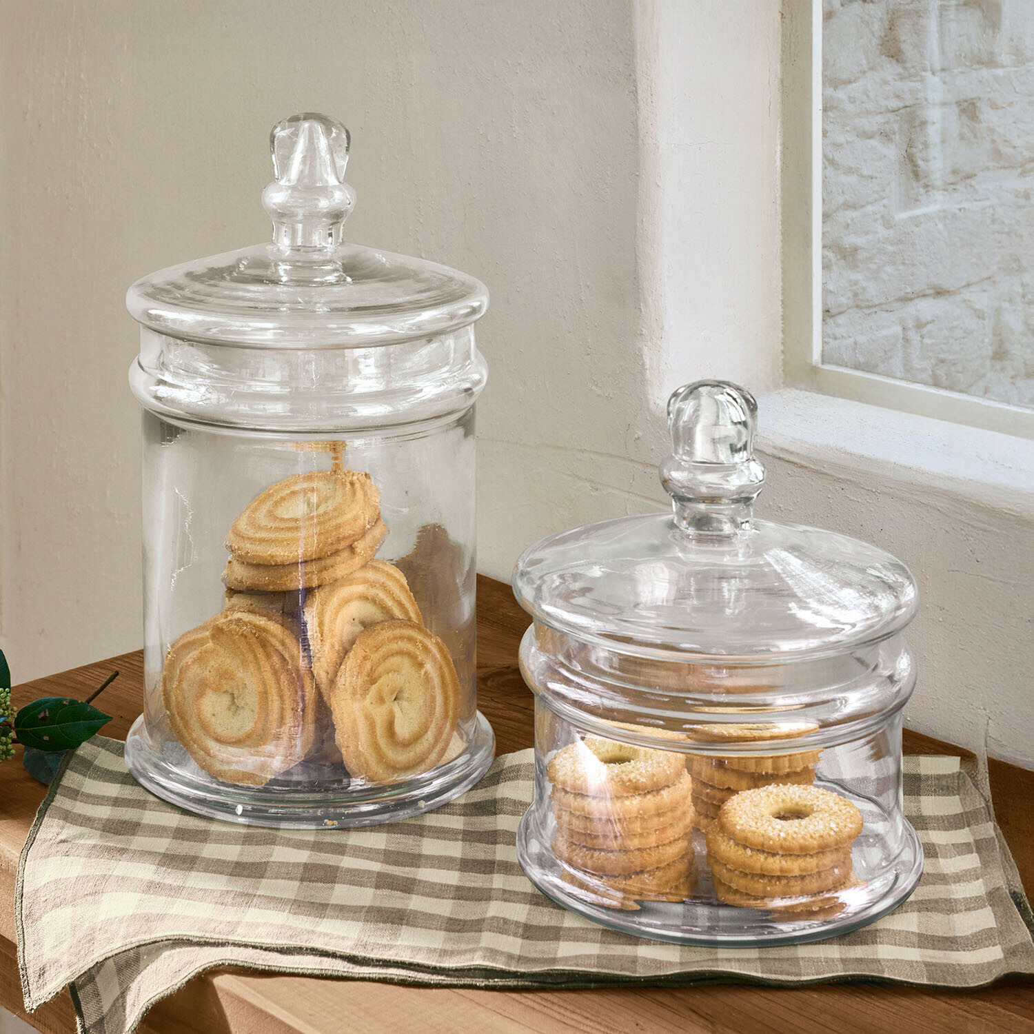 Zwei transparente Glasbehälter mit Deckeln und Keksen auf kariertem Tuch auf Holztisch