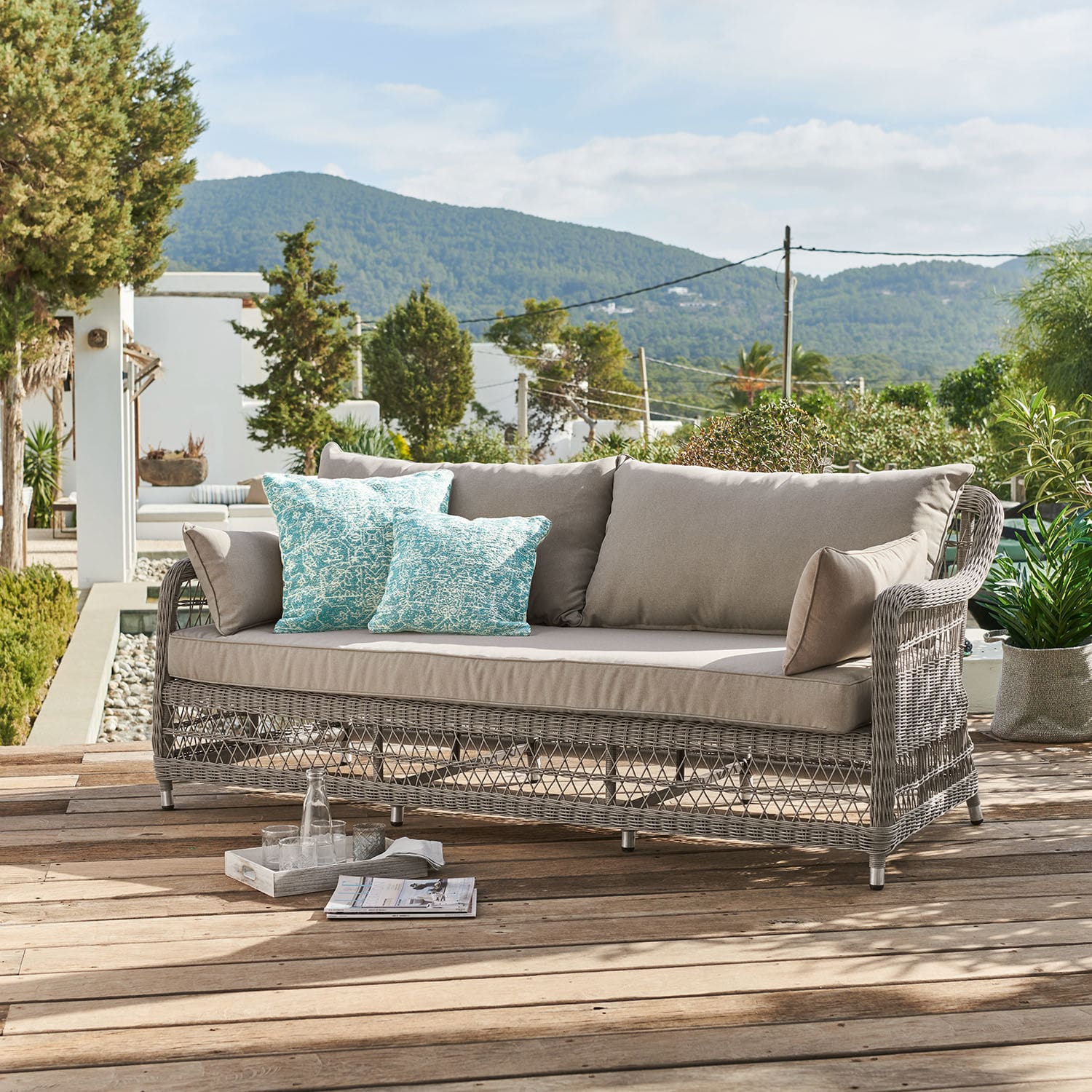 Sofa Tanaiste Gartensofa aus grauem Rattan mit beigen Polstern und türkisfarbenen Dekokissen auf Holzterrasse