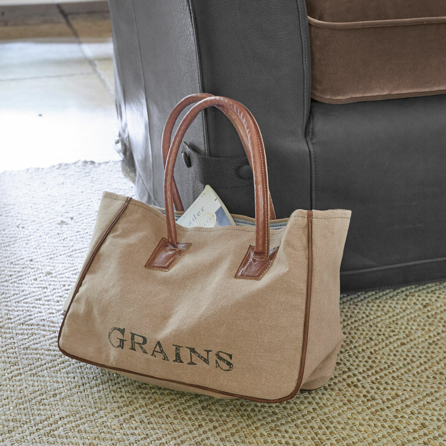 Tasche Grains Beige Stofftasche mit braunen Ledergriffen und dem Schriftzug GRAINS auf dem Teppich neben dem Sofa