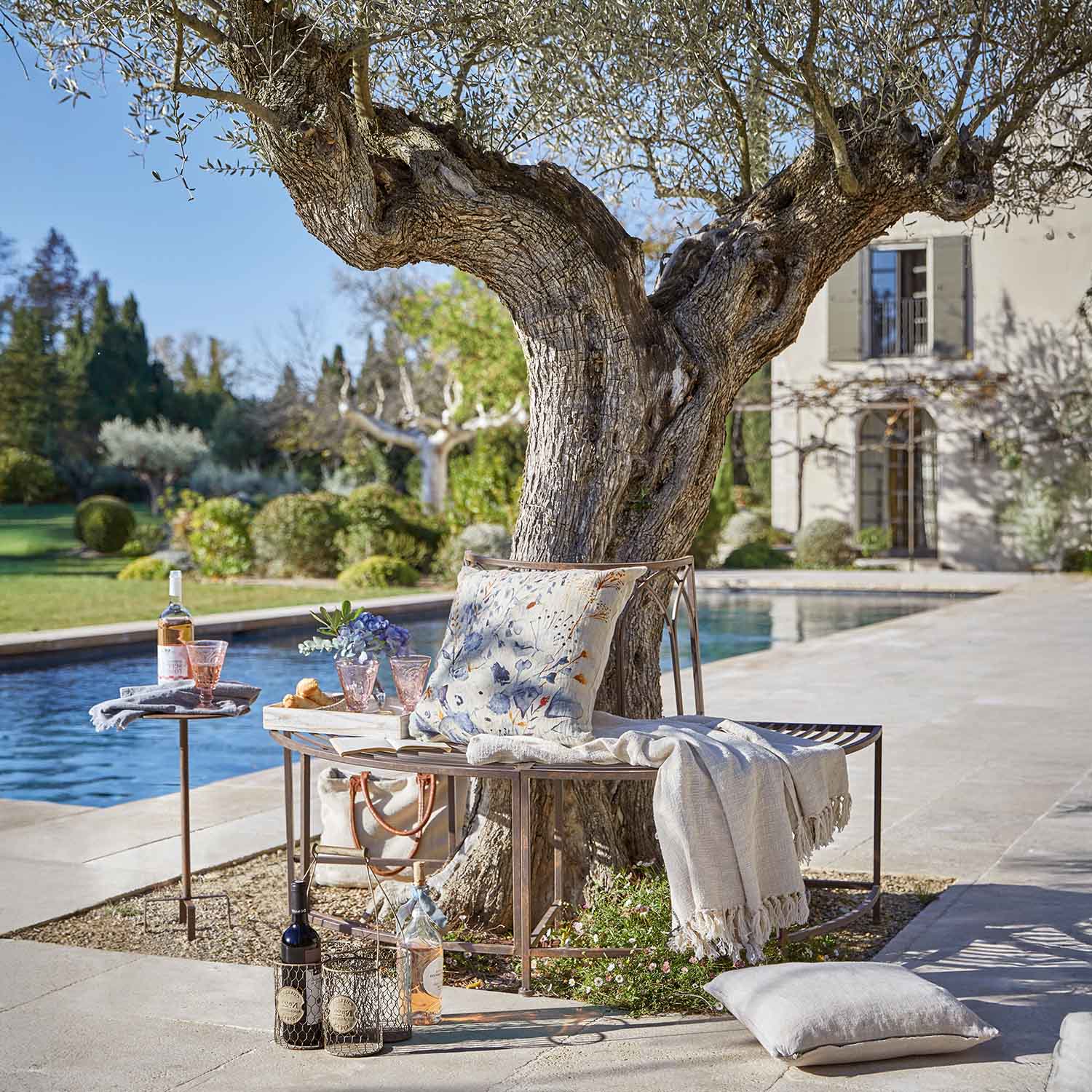 Metallbank um Baum mit Kissen und Decke am Pool im Garten mit Weinflaschen