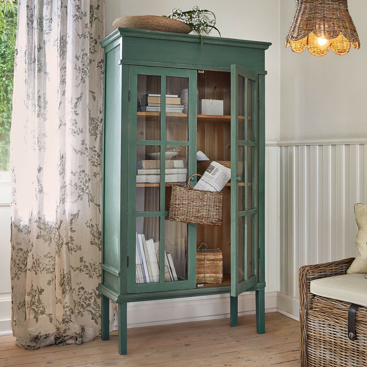 Grüner Holzvitrinenschrank mit Glastüren und Weidenkorb im hellen Wohnzimmer mit natürlichem Licht