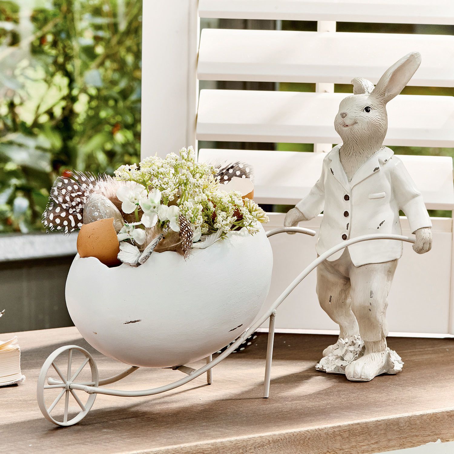 Hase Jaris Dekorativer weisser Hase mit Keramikei und Blumen in Metallwagen auf dem Tisch.